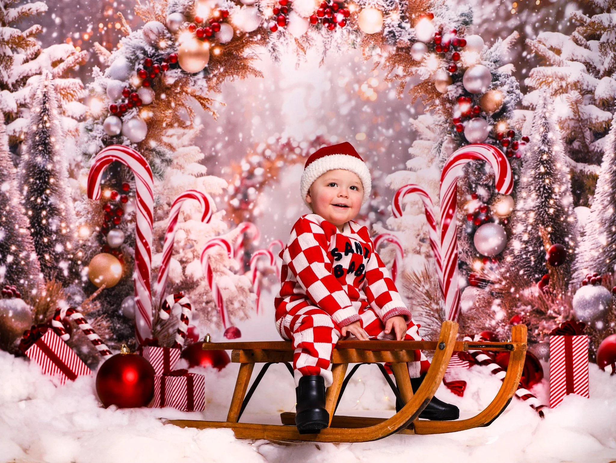 A young child dressed in red and white checkered pajamas and a Santa hat sits on a wooden sled surrounded by Christmas decorations, candy canes, ornaments, and a snowy festive backdrop.