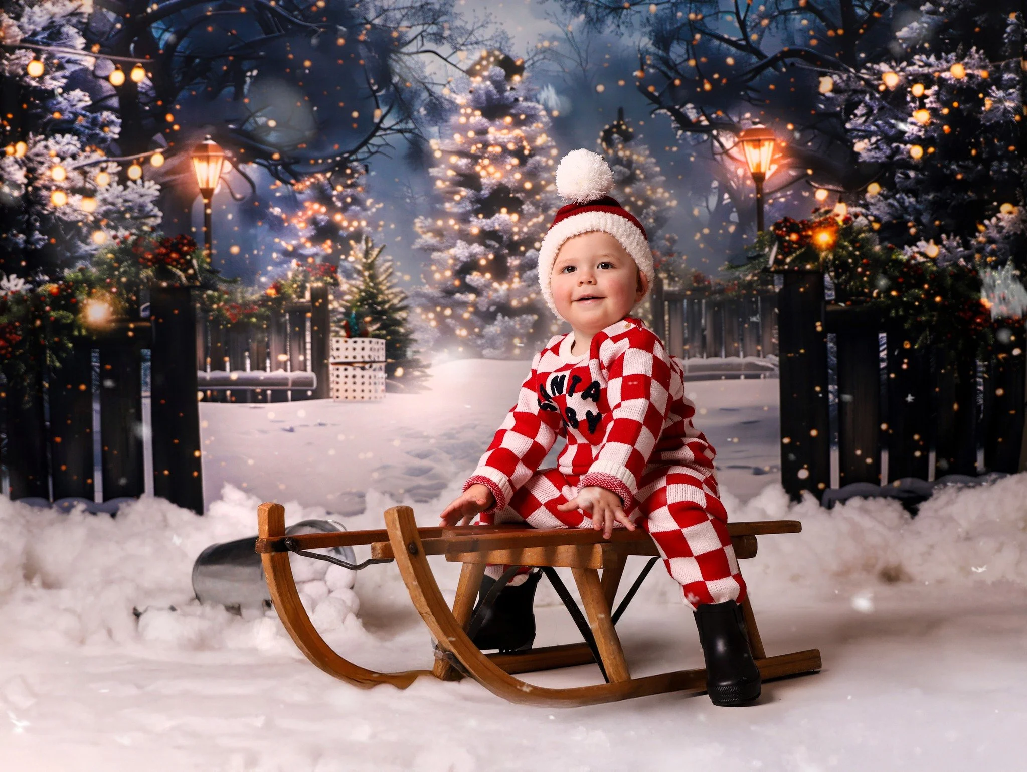 A smiling baby in holiday pajamas and a Santa hat sitting on a wooden sled in a snowy winter scene with Christmas trees and lit lamps.