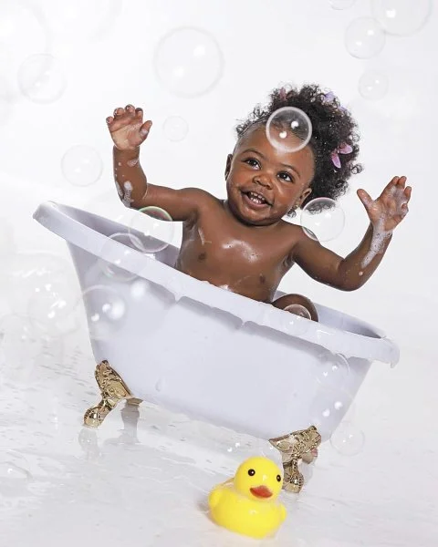 A smiling toddler with curly hair and a pink bow sitting in a white bathtub surrounded by bubbles, with a yellow rubber duck in front from their cake smash photoshoot at Meadowside Studio in Warrington to celebrate their 1st birthday!