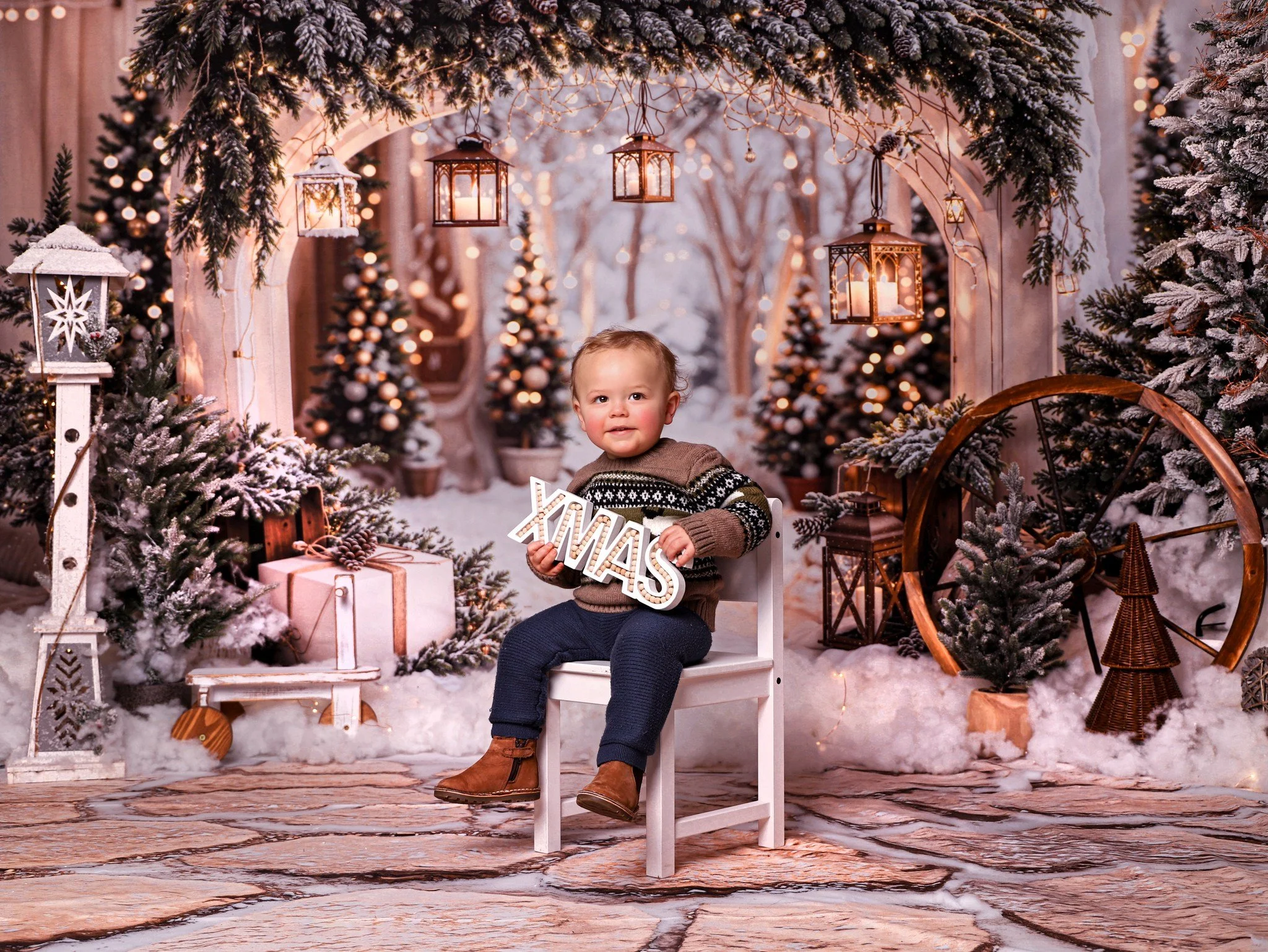A young child sitting on a white chair in a Christmas winter scene with snow-covered trees, wrapped gifts, and festive lanterns hanging from an arch.