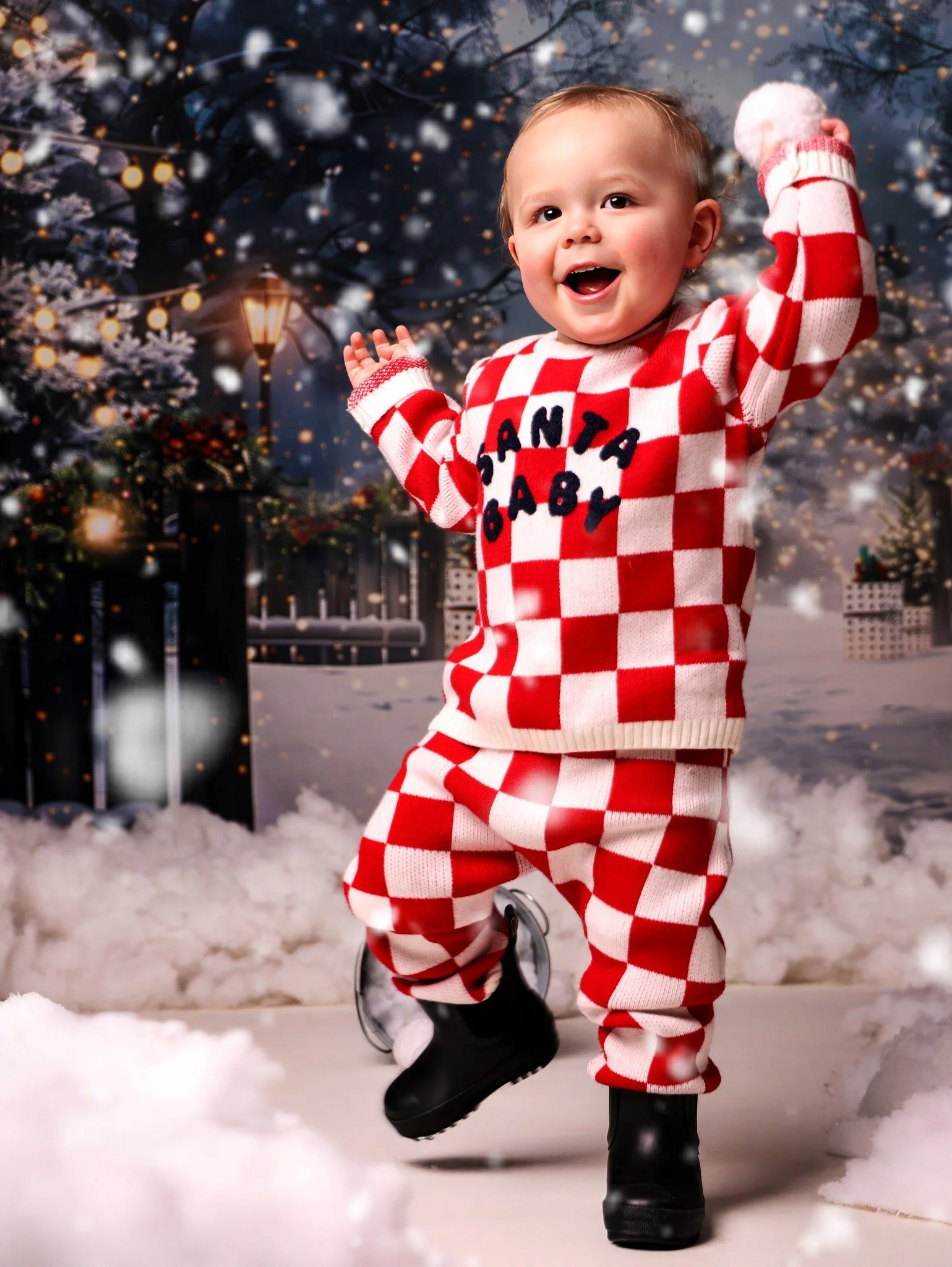 A young child in winter clothing, wearing a red and white checkered sweater and pants, black boots, and a pink pom-pom hat, is playing in snow outdoors during winter with festive lights and decorated trees in the background.