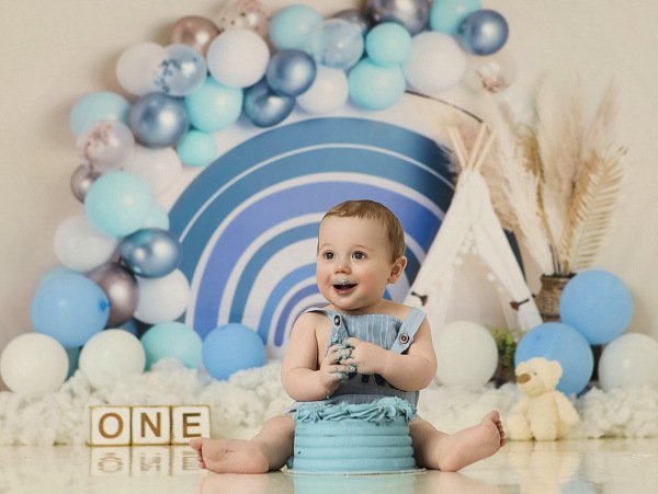 A smiling baby sitting on the floor in front of a blue and white balloon arch, with a small cream-colored stuffed animal and blocks spelling "ONE" nearby, celebrating a first birthday at Meadowside studio in Warrington UK