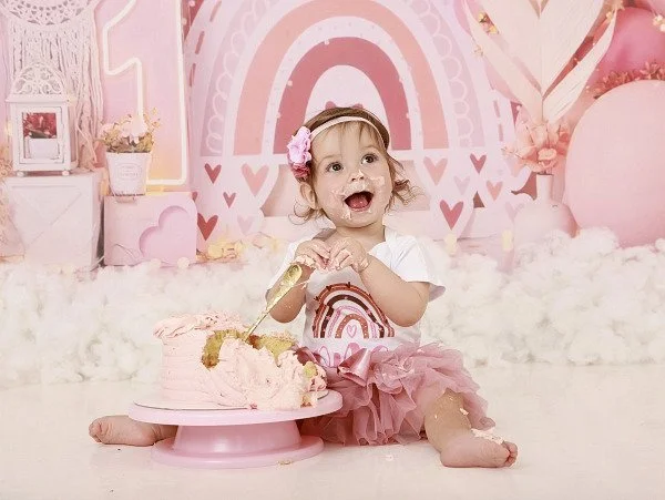A young girl celebrating a birthday with a pink cake, sitting on a fluffy white rug in a pink-themed decorated room from their cake smash photoshoot at Meadowside Studio in Warrington to celebrate their 1st birthday!