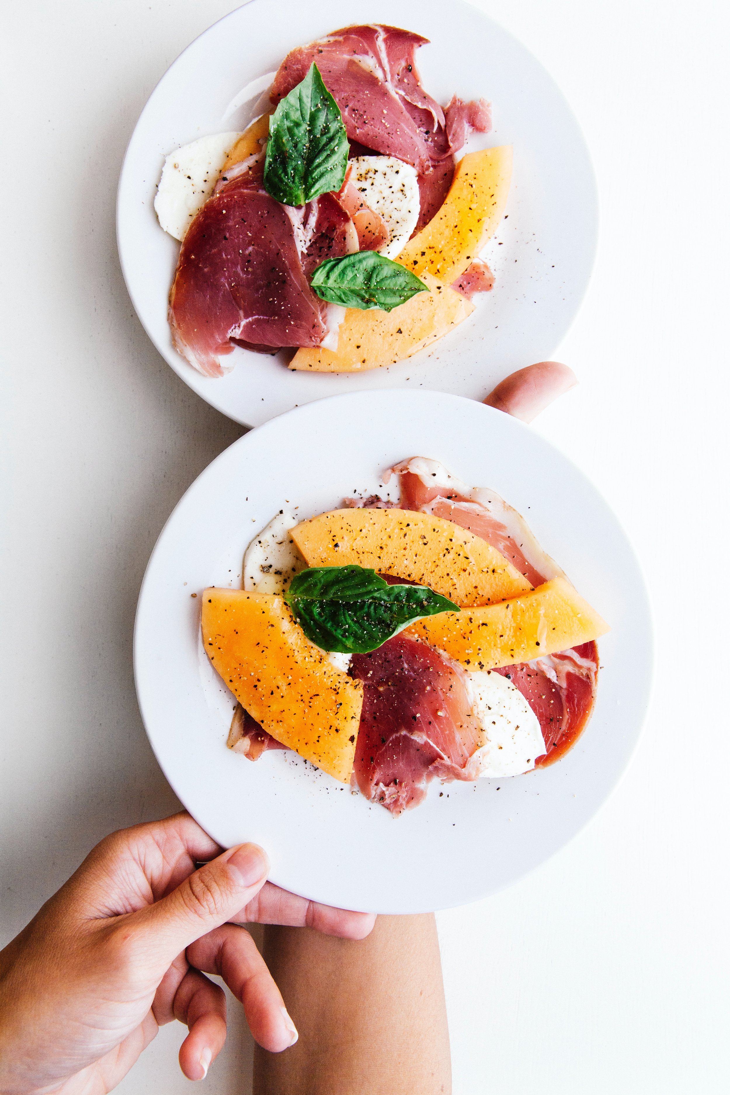 Two plates of Italian antipasto with prosciutto, melon slices, mozzarella, and basil leaves.