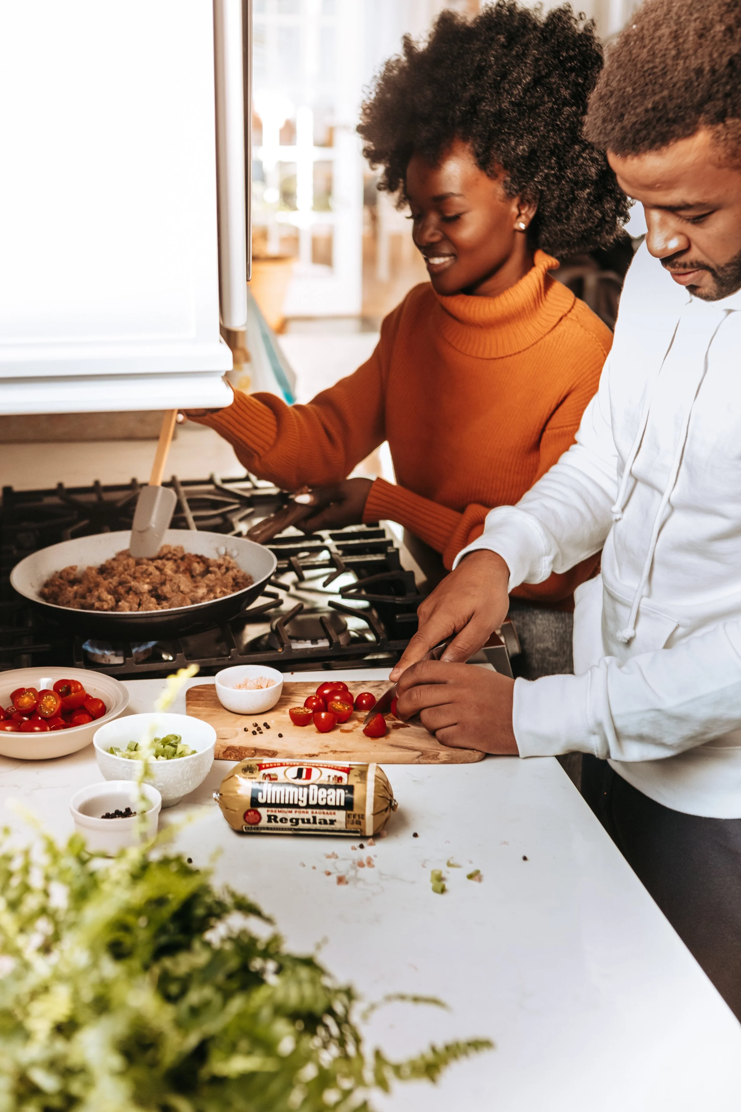 Two people cooking in a kitchen, chopping tomatoes and cooking sausage on a stove.