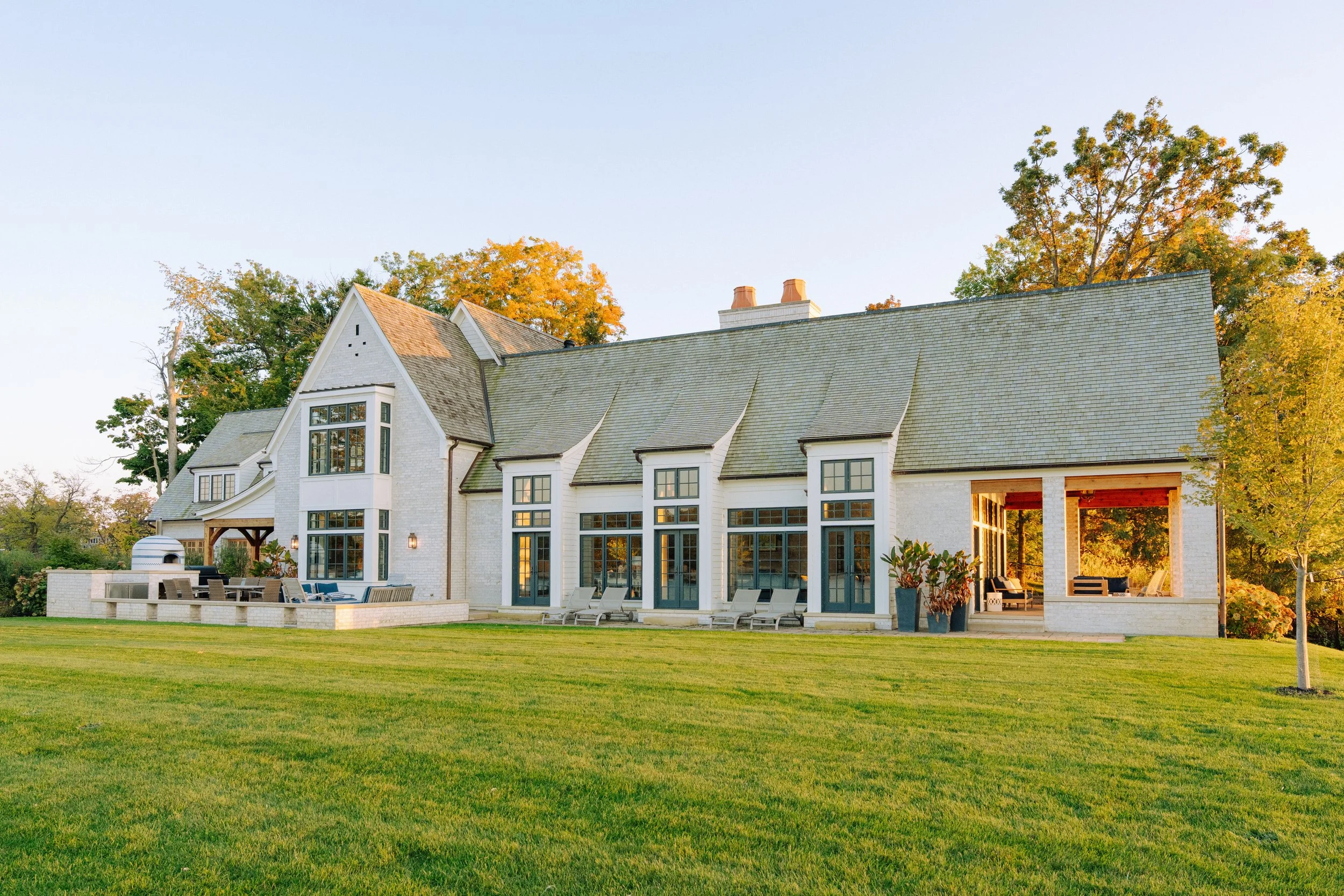 A large modern house with a well-maintained lawn in front, featuring white exterior walls, multiple large windows, and a covered patio area with outdoor furniture, set against a backdrop of trees with some orange and green leaves.