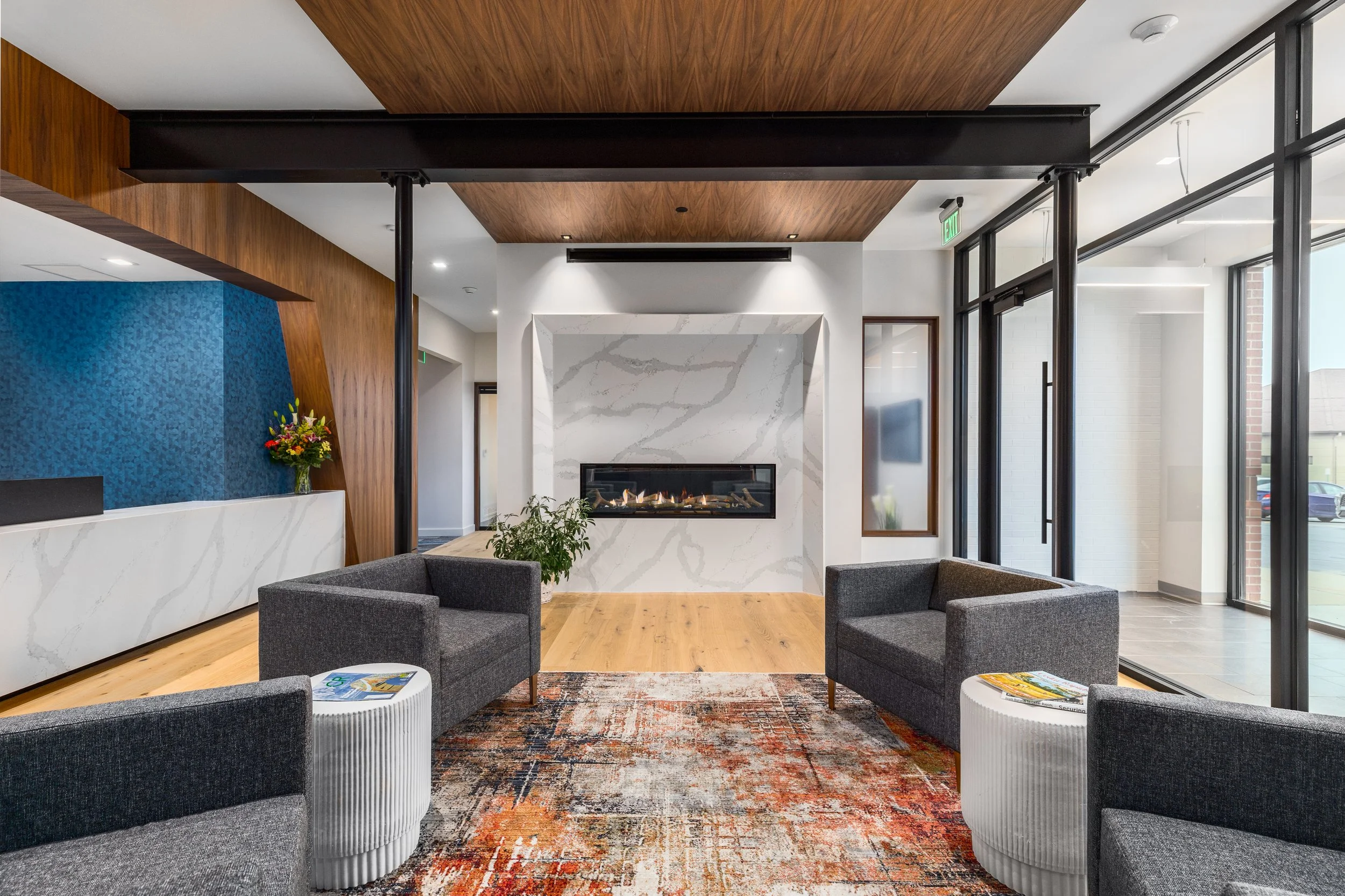 A glimpse into the Carson Wealth offices - a modern lobby with a marble fireplace, four dark gray armchairs, white side tables with magazines, a colorful rug, and large glass windows and doors showcasing services provided by Form Function Studio.