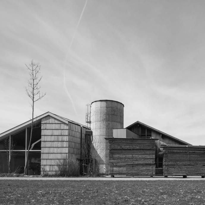 Black and white photo of a modern barn or farmhouse with a silo, a leafless tree, and a clear sky with contrails.