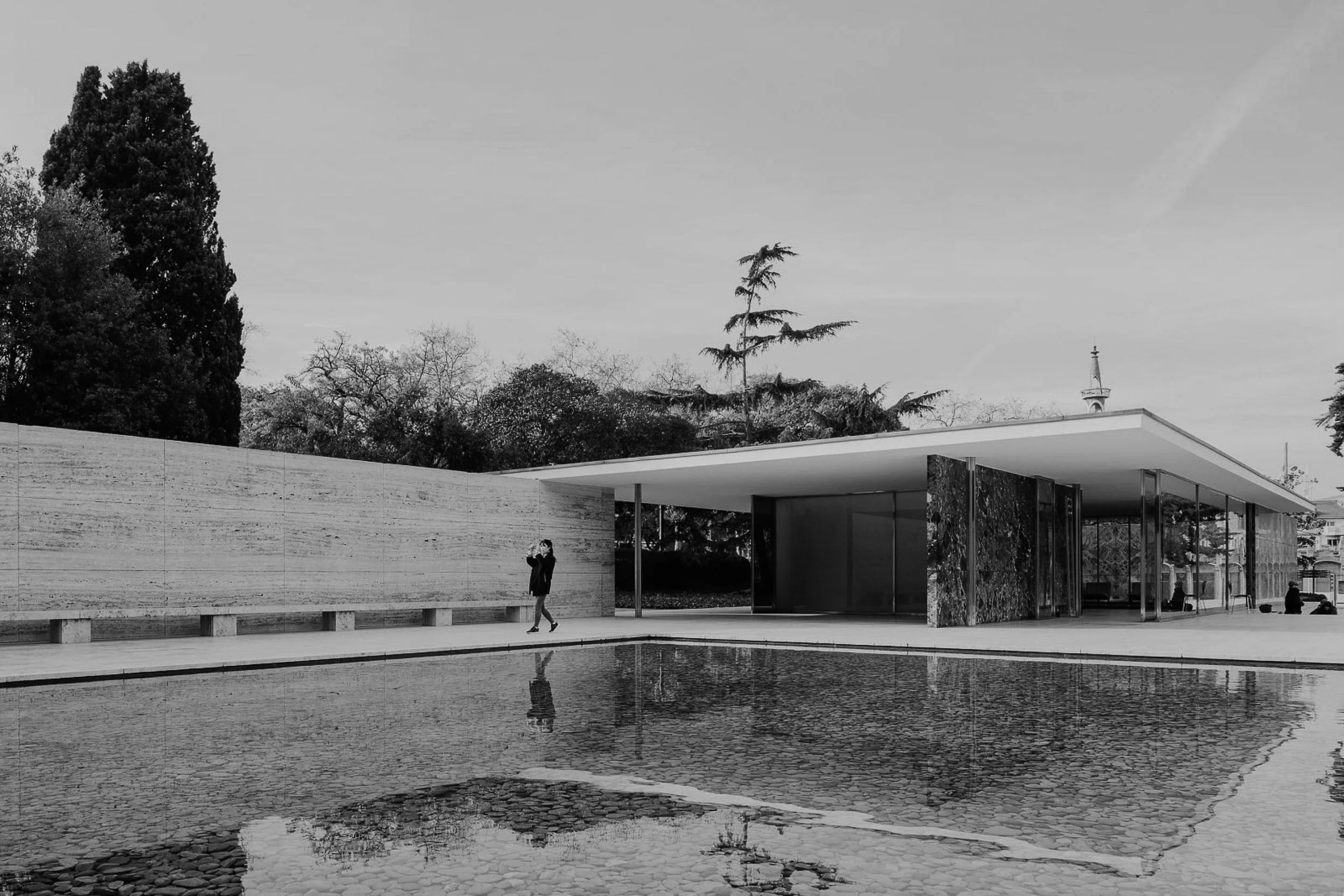 A modern building with a flat roof next to a shallow reflective pool, with a person walking along the pathway and trees in the background.