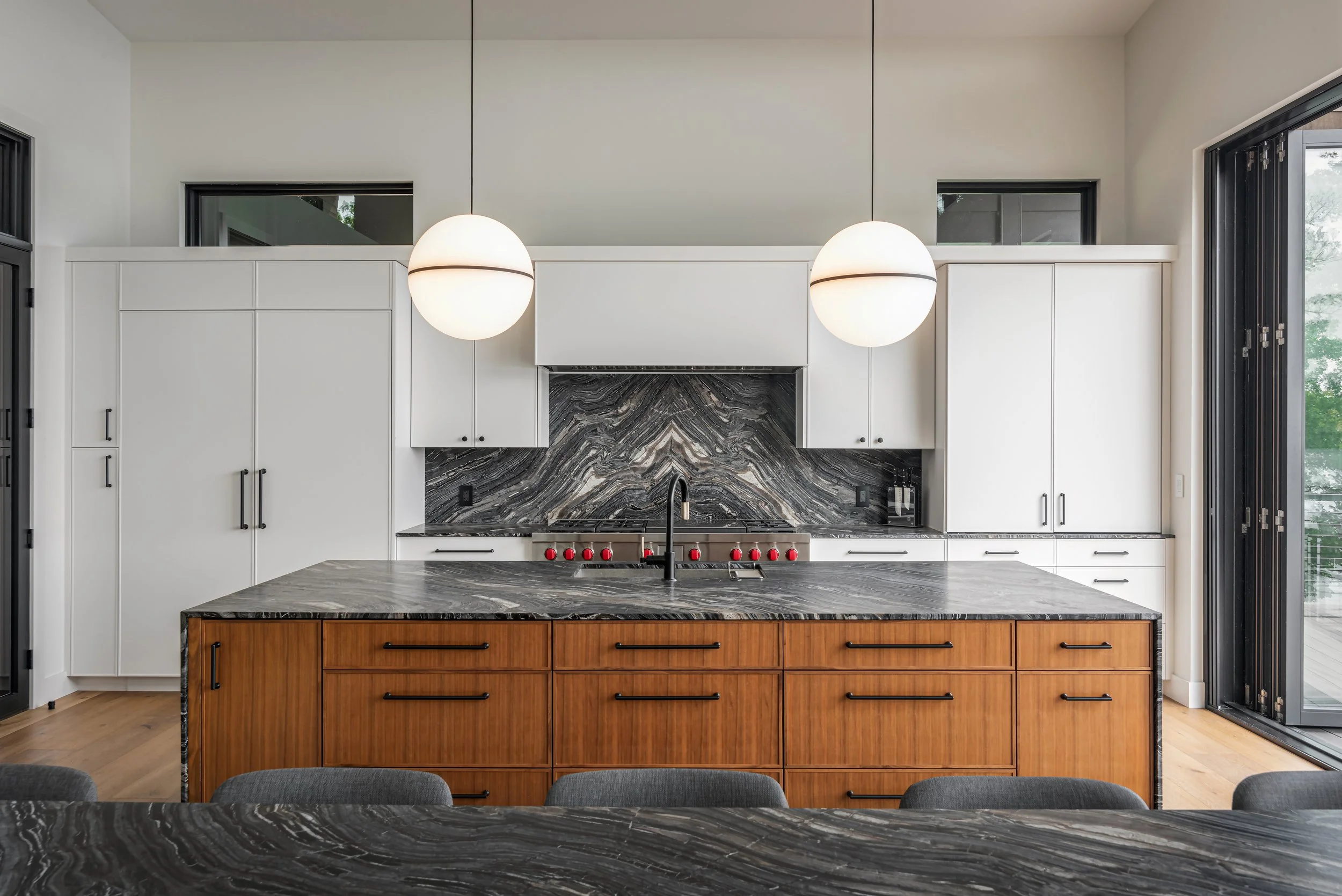 Modern kitchen with white cabinetry, black marble backsplash, and a wooden island with a marbled countertop. Two large white pendant lights hang above the island, and sliding glass doors are visible on the right.