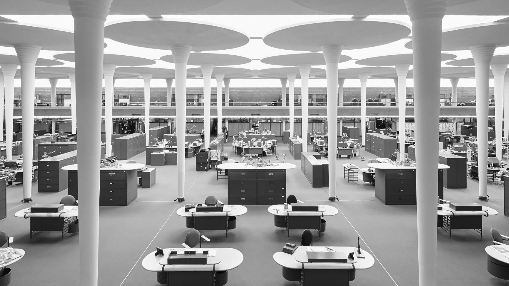 An empty open office space with multiple desks, chairs, and filing cabinets, characterized by tall white structural columns and a circular ceiling design.
