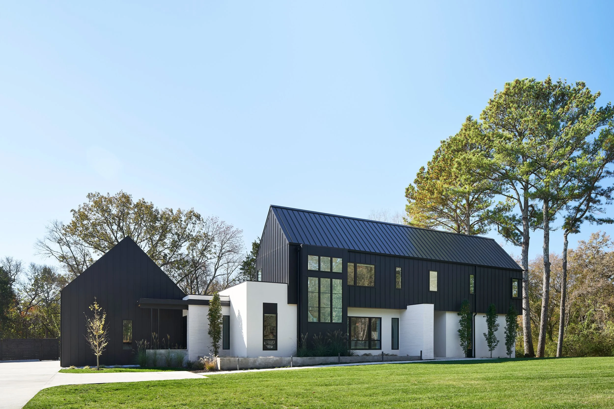 Modern house with black and white exterior, large windows, and a well-maintained lawn under a clear blue sky, surrounded by tall trees.