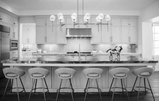 Modern kitchen with white cabinets, a large island with six bar stools, hanging light fixtures, and a flower arrangement on the countertop.