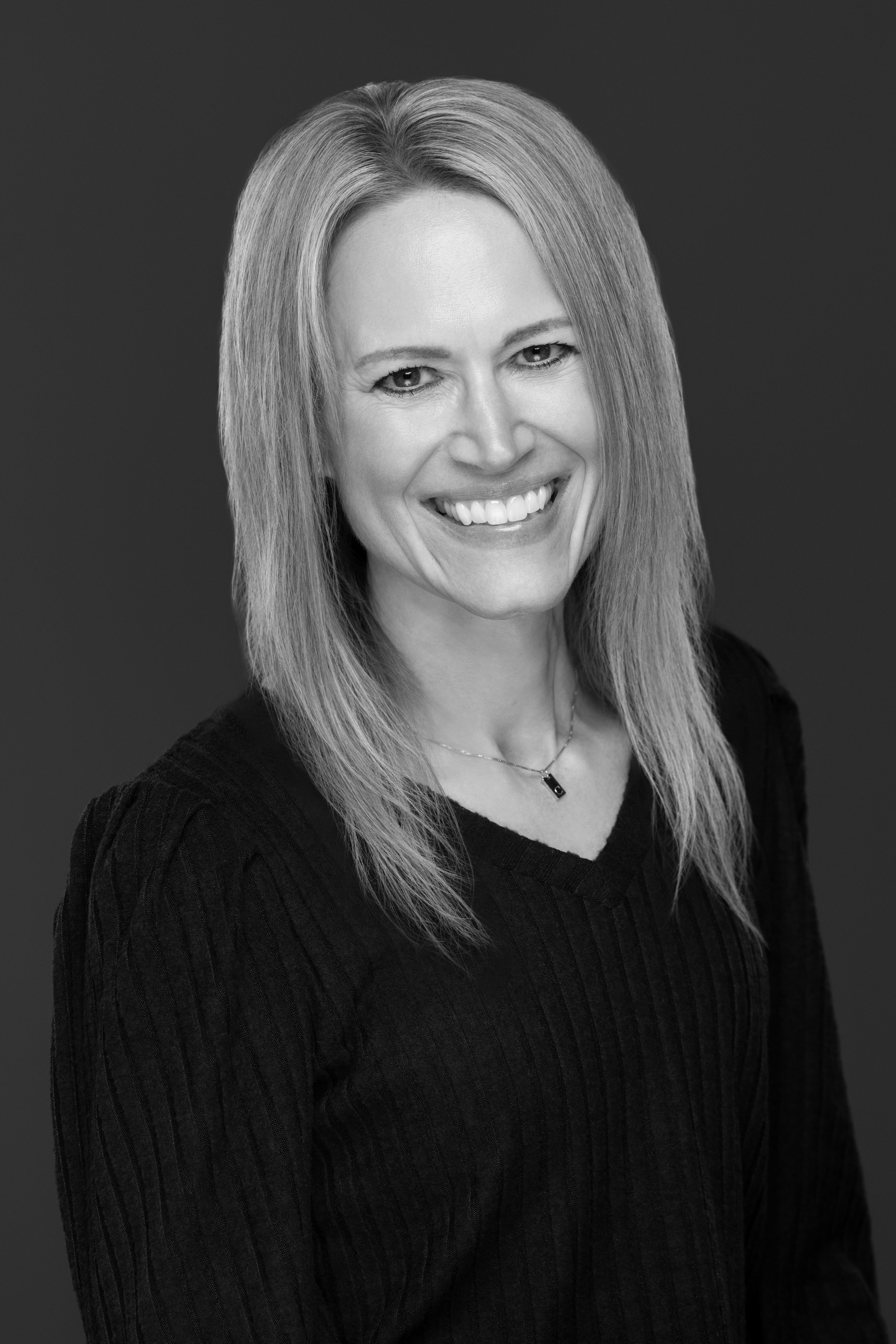 Black and white portrait of a smiling woman with straight, shoulder-length hair, wearing a dark, ribbed top and a necklace, against a plain background.