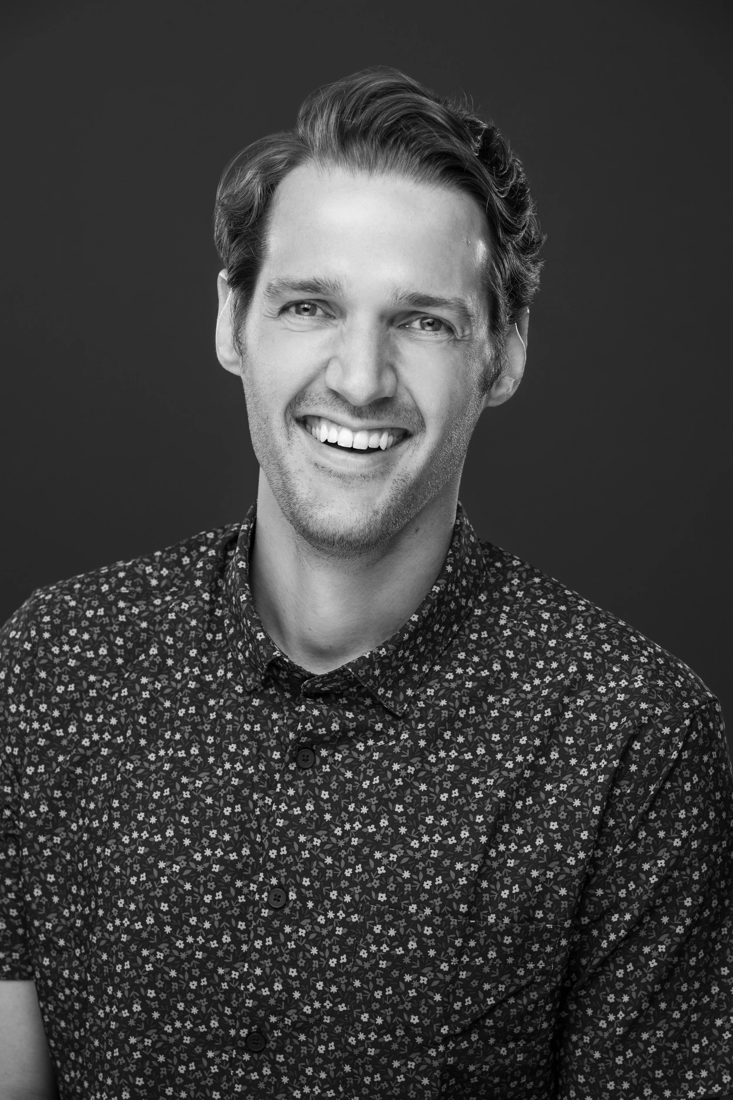 Black and white portrait of a smiling man with wavy hair, wearing a collared button-up shirt with a floral pattern, against a dark background.