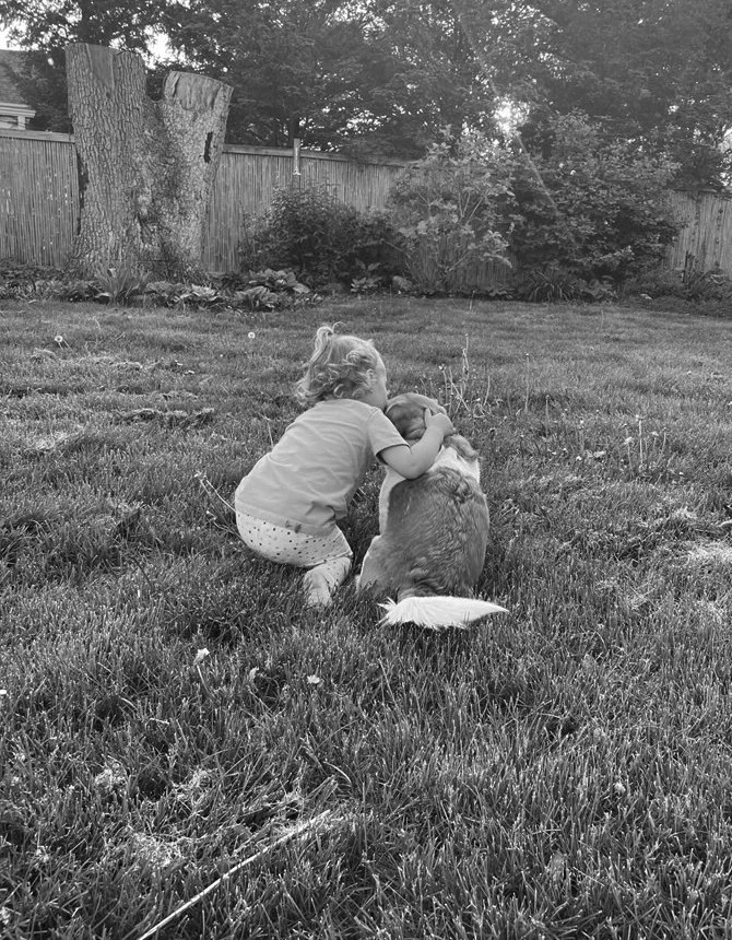 A young girl is kneeling on grass, kissing a dog on the head in a backyard with trees, bushes, a wooden fence, and a large tree stump in the background.