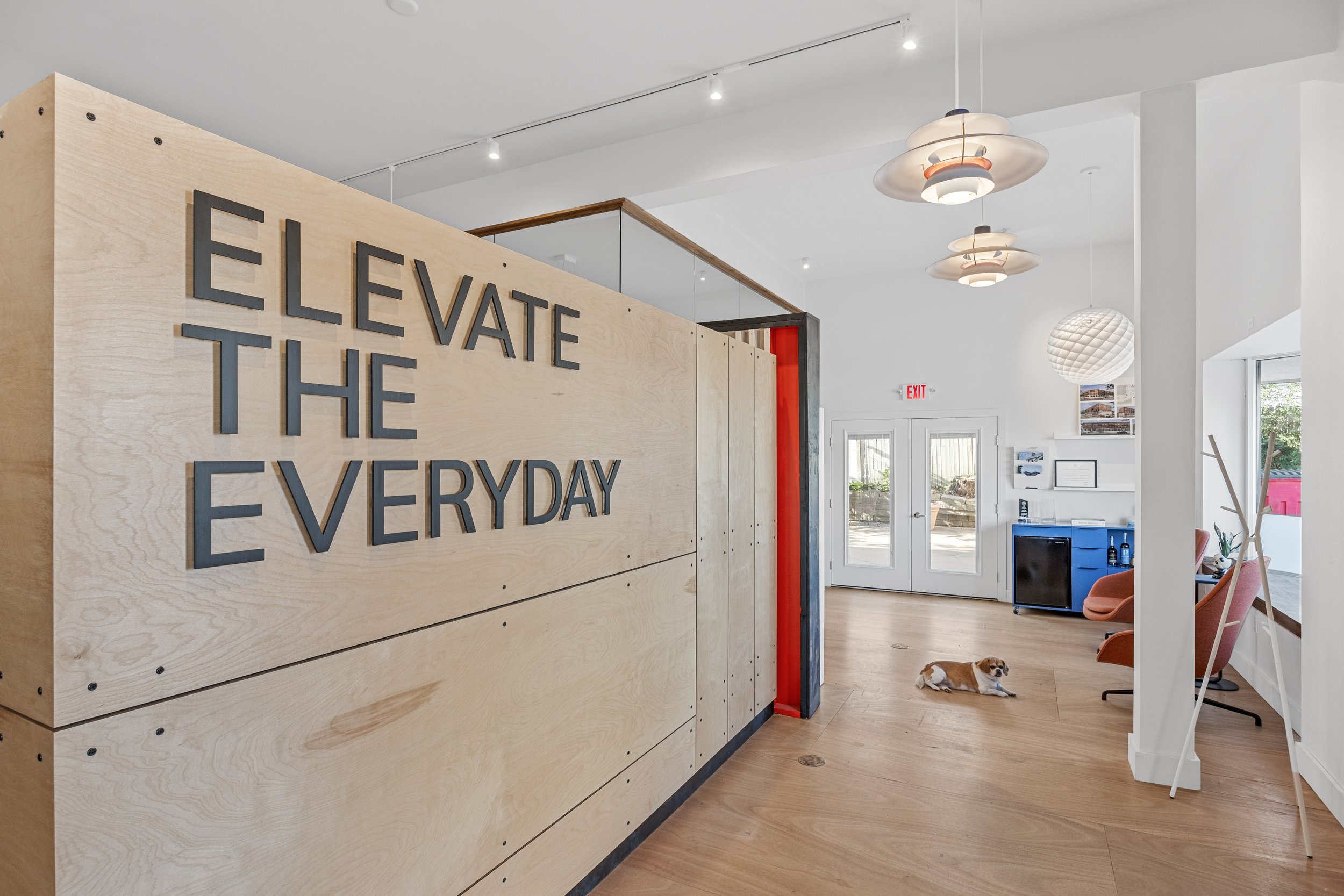 Modern interior reception area with a wooden sign that reads "Elevate the Everyday." There are white walls, hardwood floors, pendant lighting, a sitting area with pink and orange chairs, a small blue cabinet, and a dog lying on the floor near the entrance doors.