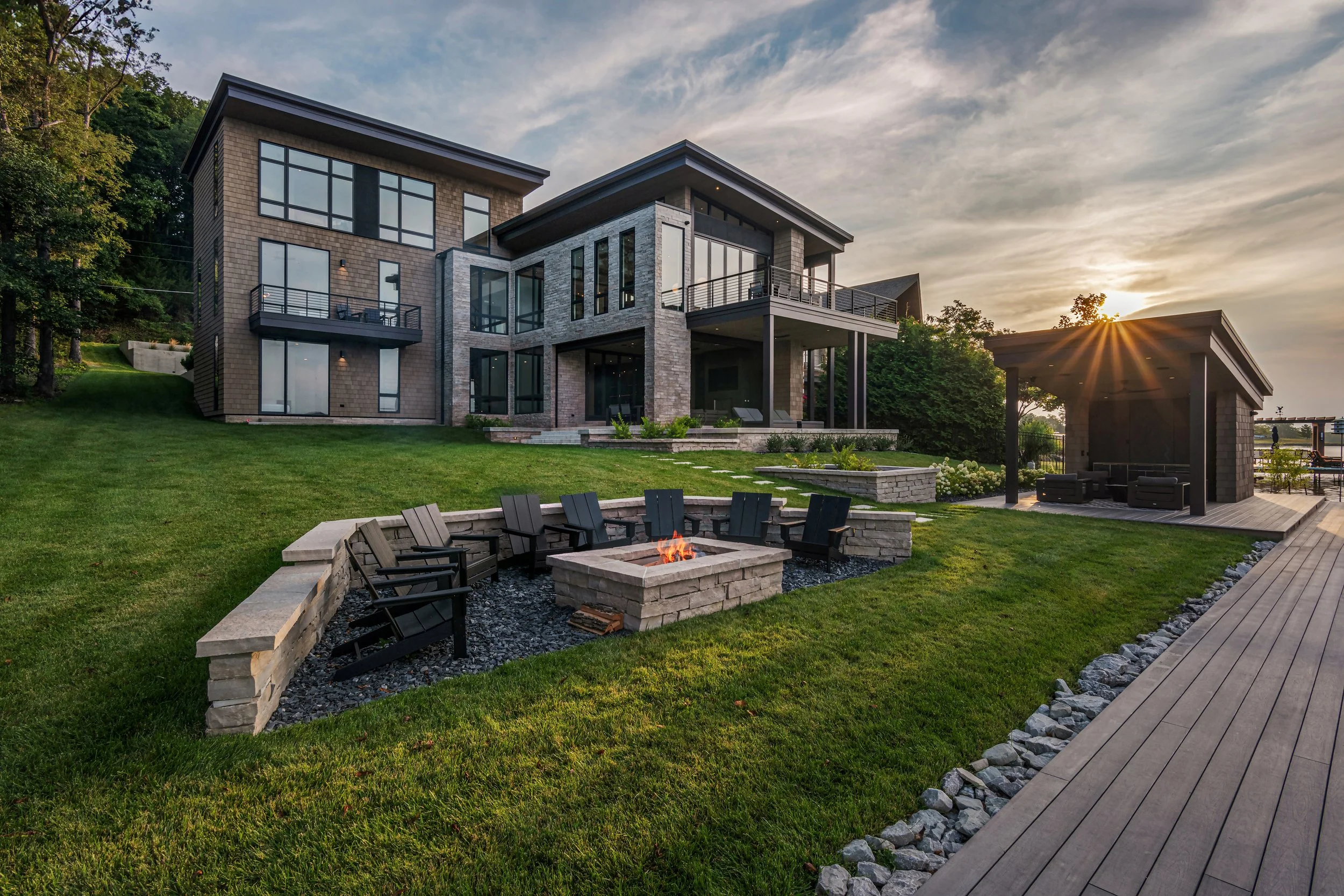 Modern multi-story house with large windows, a backyard with a fire pit surrounded by chairs, a raised deck, and a gazebo at sunset.