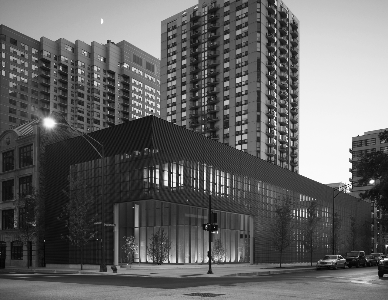 Black and white photo of a city street with a modern building in the foreground and high-rise apartment buildings in the background. The street has parked cars and streetlights, with a crescent moon in the sky.