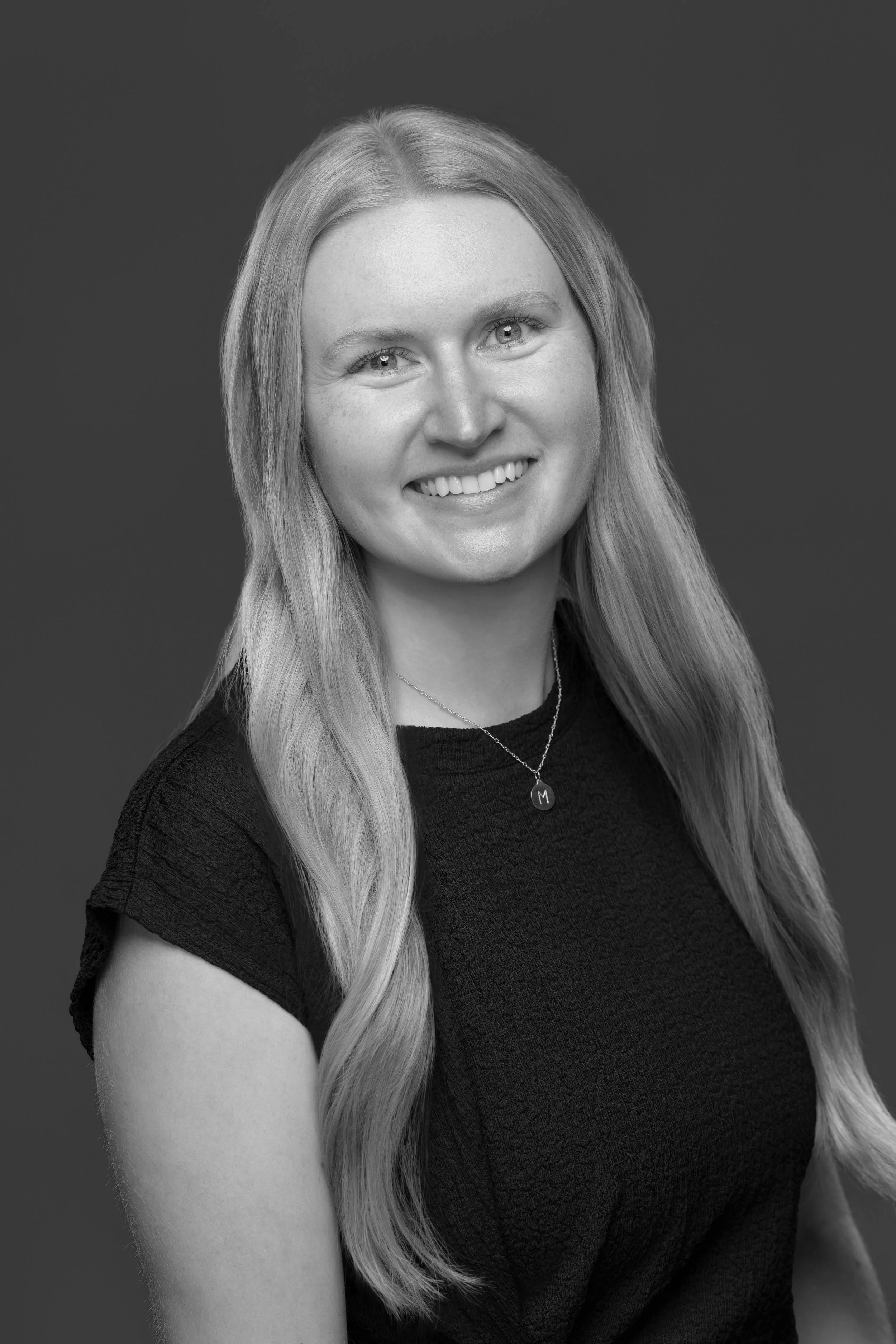 Black and white portrait of a smiling woman with long wavy hair, wearing a dark shirt and a necklace with a letter 'M' pendant.