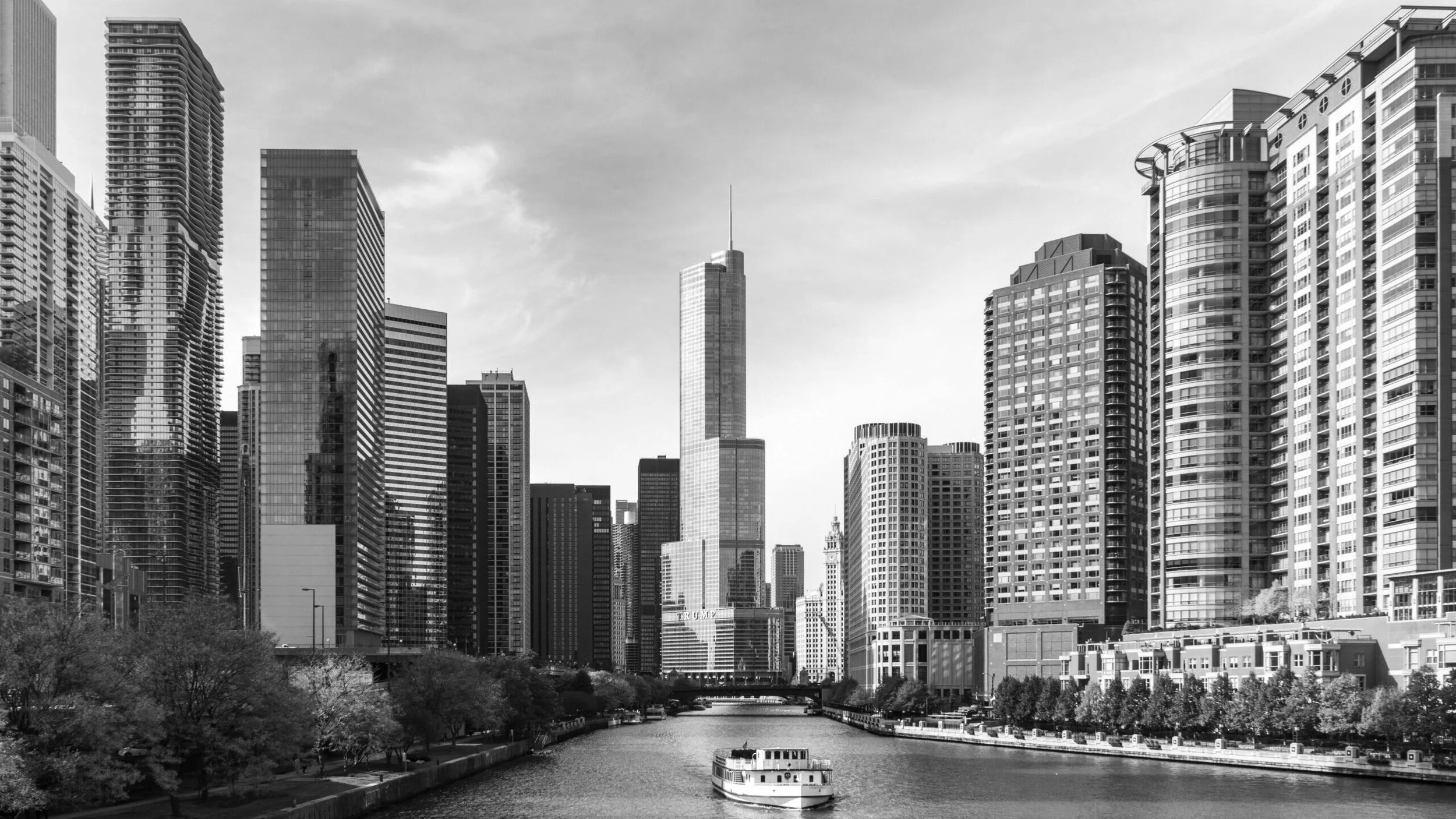 Black and white photo of downtown Chicago skyline with tall buildings and a river in the foreground, with a boat on the river and trees along the riverbank.