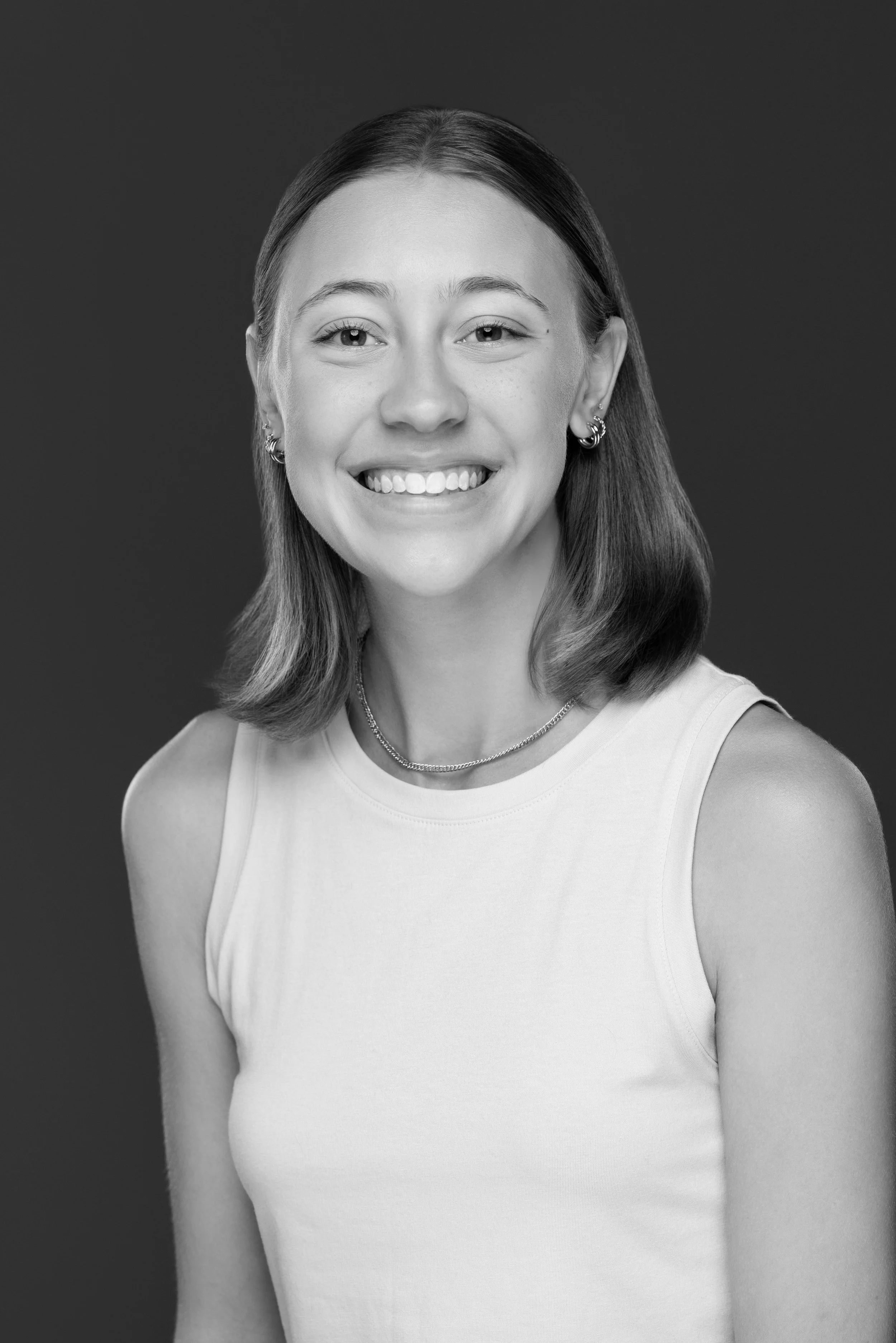 A smiling young woman with shoulder-length hair, wearing a sleeveless top, earrings, and a necklace, against a plain background.