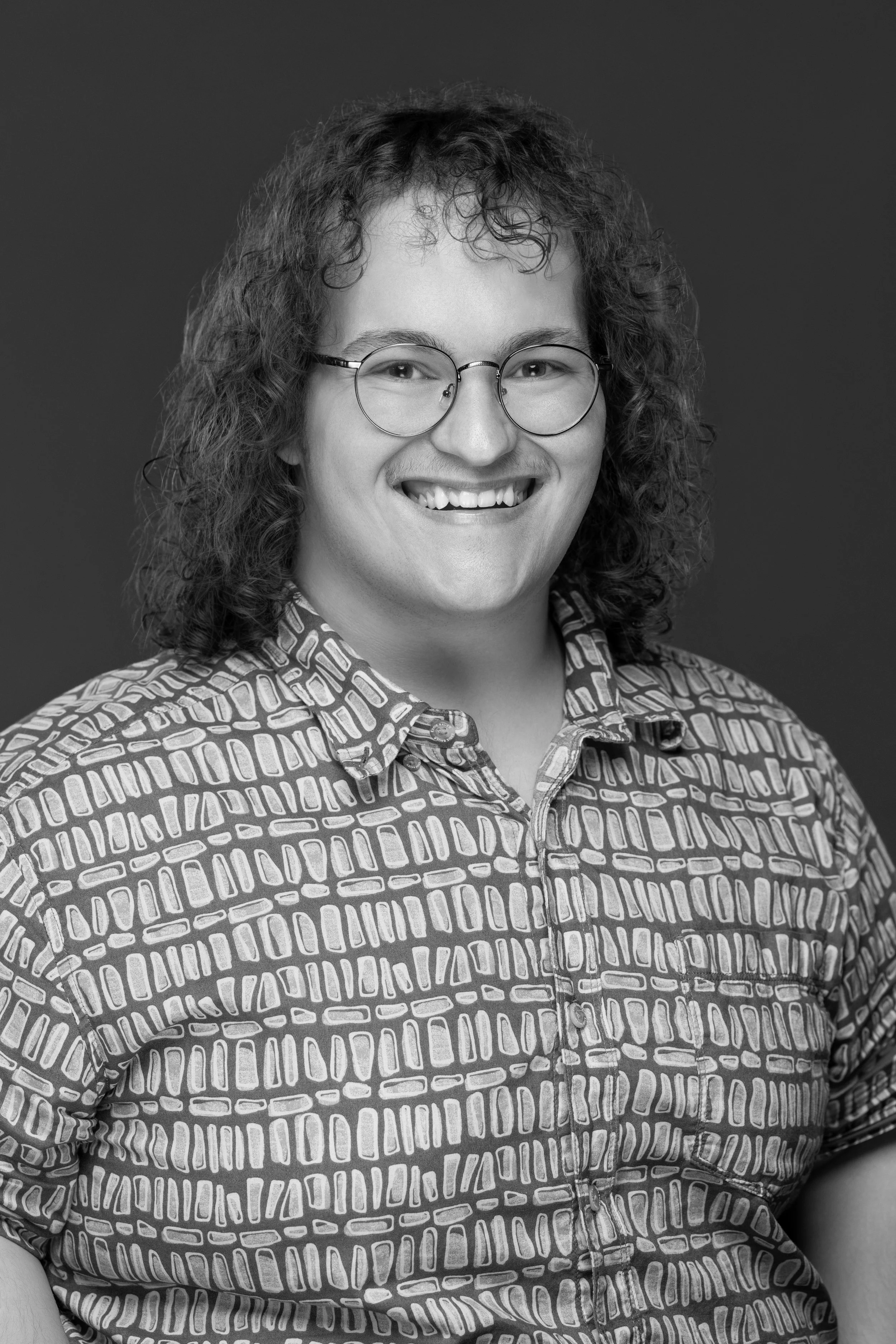 Black and white portrait of a man wearing glasses and a patterned button-up shirt, smiling at the camera.