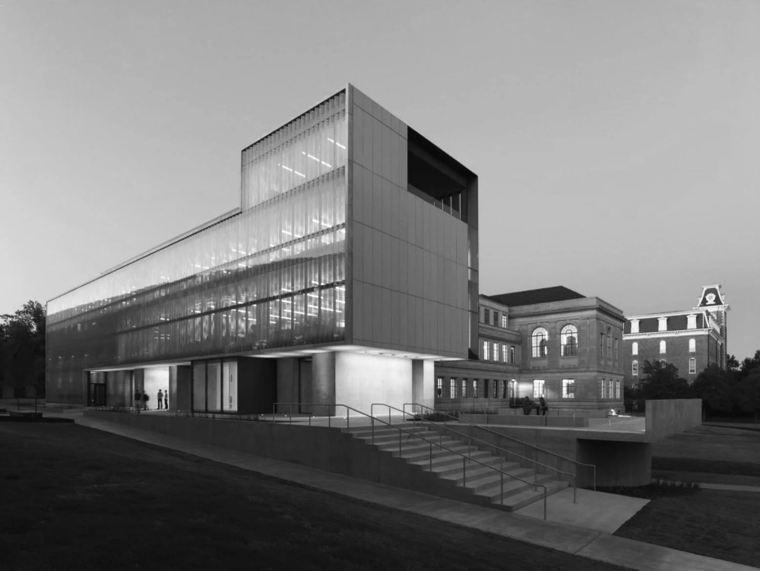 Black and white photo of a modern glass building in an urban setting, with a historical brick building in the background and a few people walking on the sidewalk.