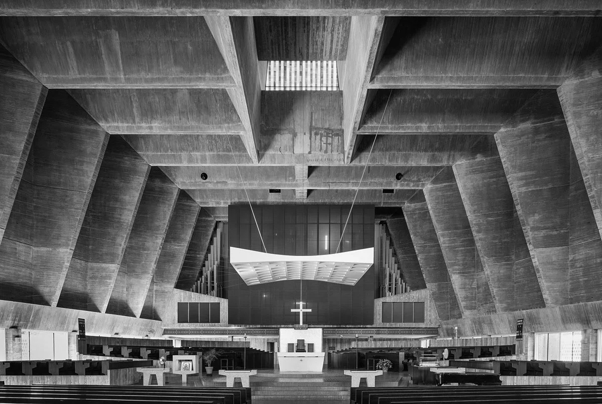 Interior view of a modern church sanctuary with a prominent cross at the altar, geometric concrete architecture, and seating arranged in front.