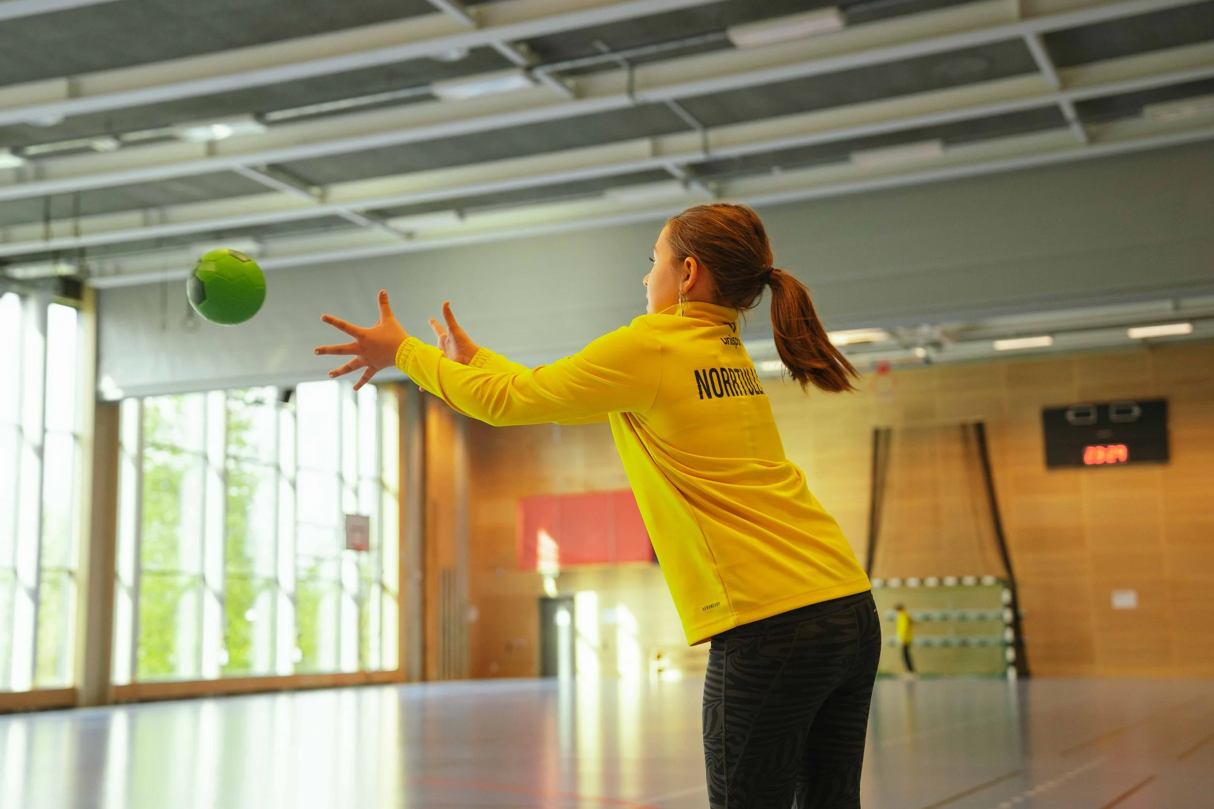 En kvinna i en gul sportjacka kastar en grön handboll inomhus i en sporthall.