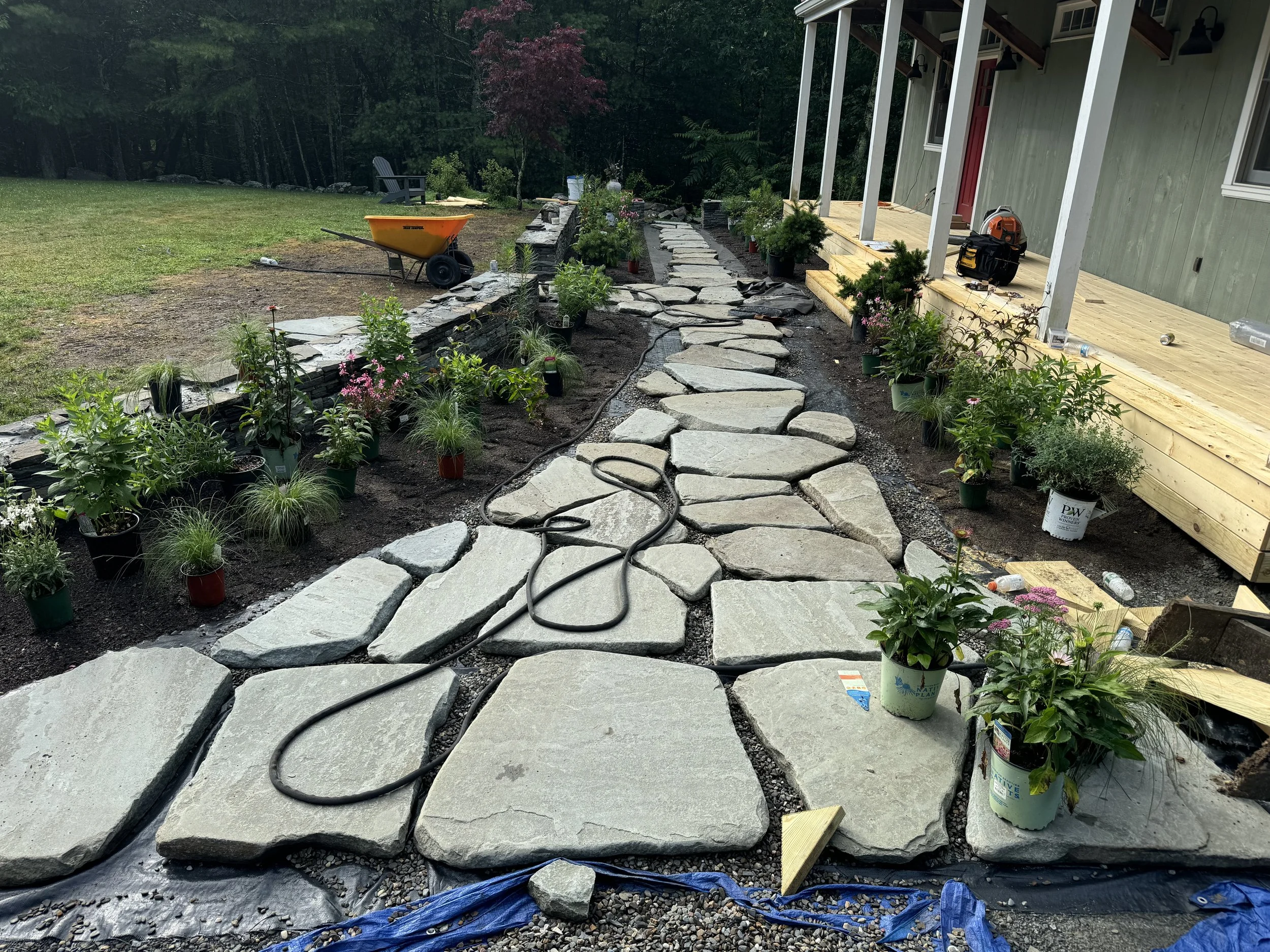 Construction scene showing a stone pathway being built in a backyard, with tools, plants, and a house on the right side.