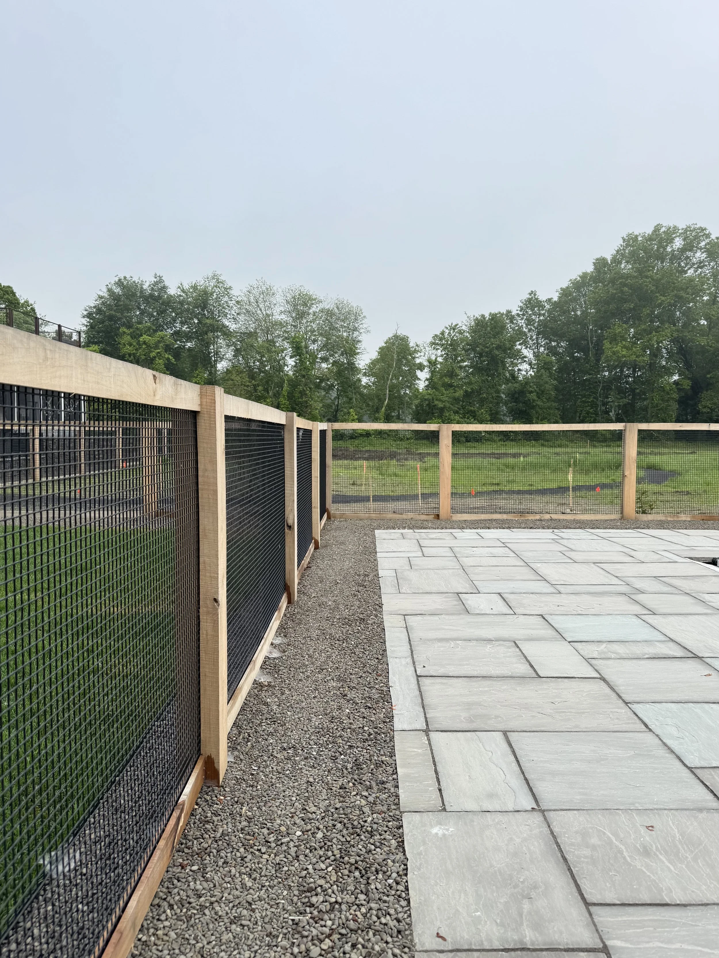 A view of a backyard with a black metal fence on the left, a wooden fence in the middle, a gravel path and a paved patio area with large gray tiles, green grass, trees, and a cloudy sky in the background.