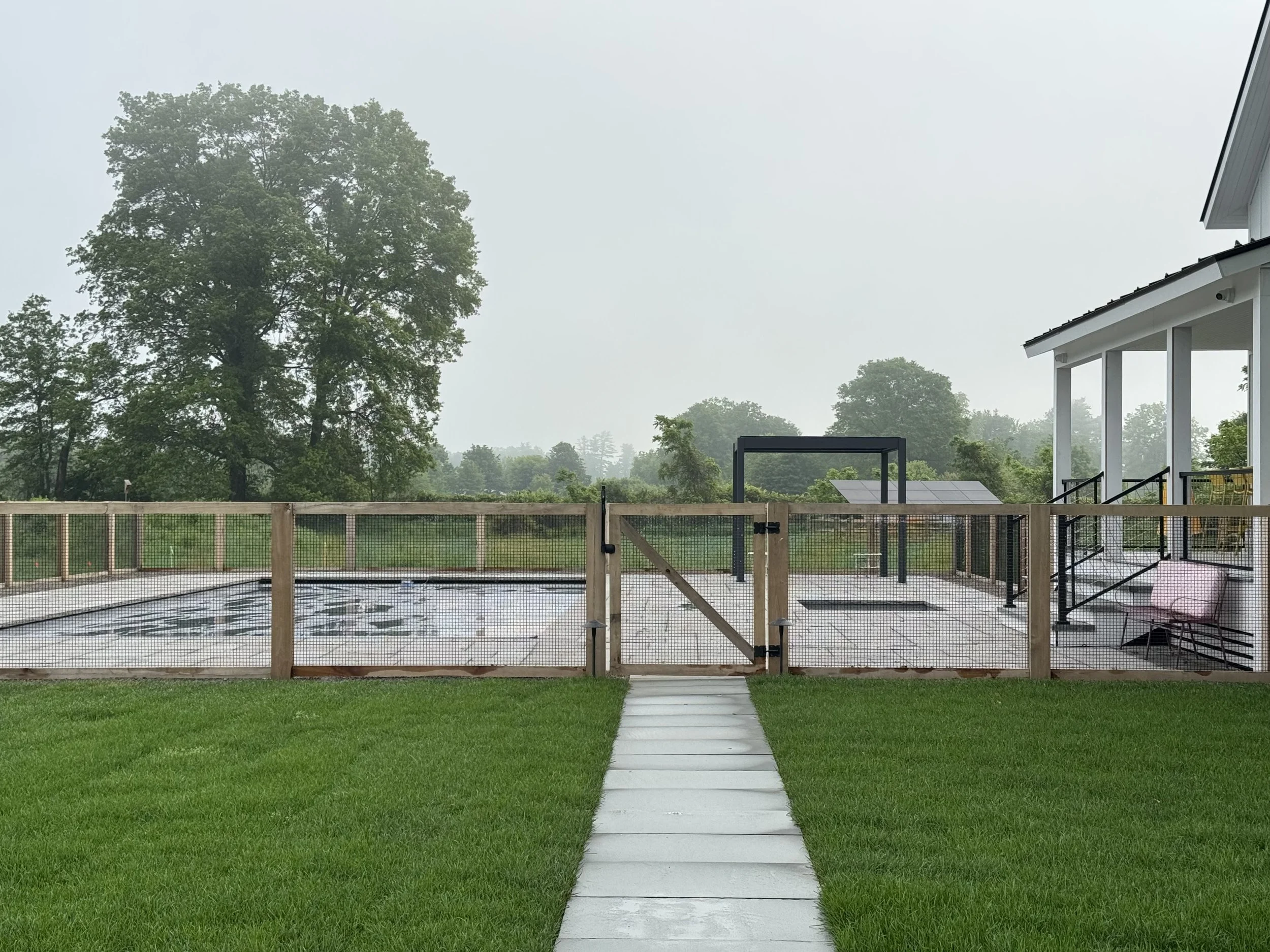 A backyard scene on a foggy day showing a fenced-in pool area with a concrete walkway, surrounded by a green lawn and trees in the background, with a white house partially visible on the right.