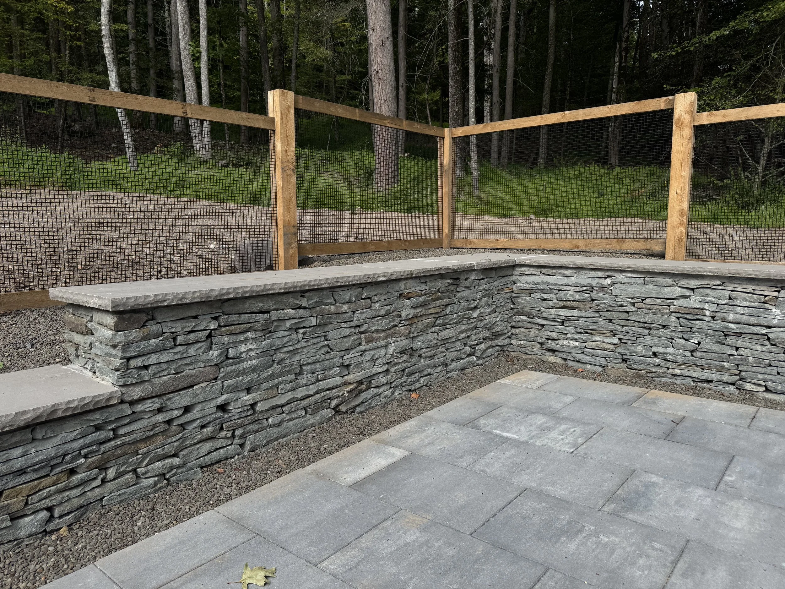 A stone retaining wall with a concrete top, gray pavers on the ground, and a wooden fence with black mesh in front of a dense forest of tall trees.