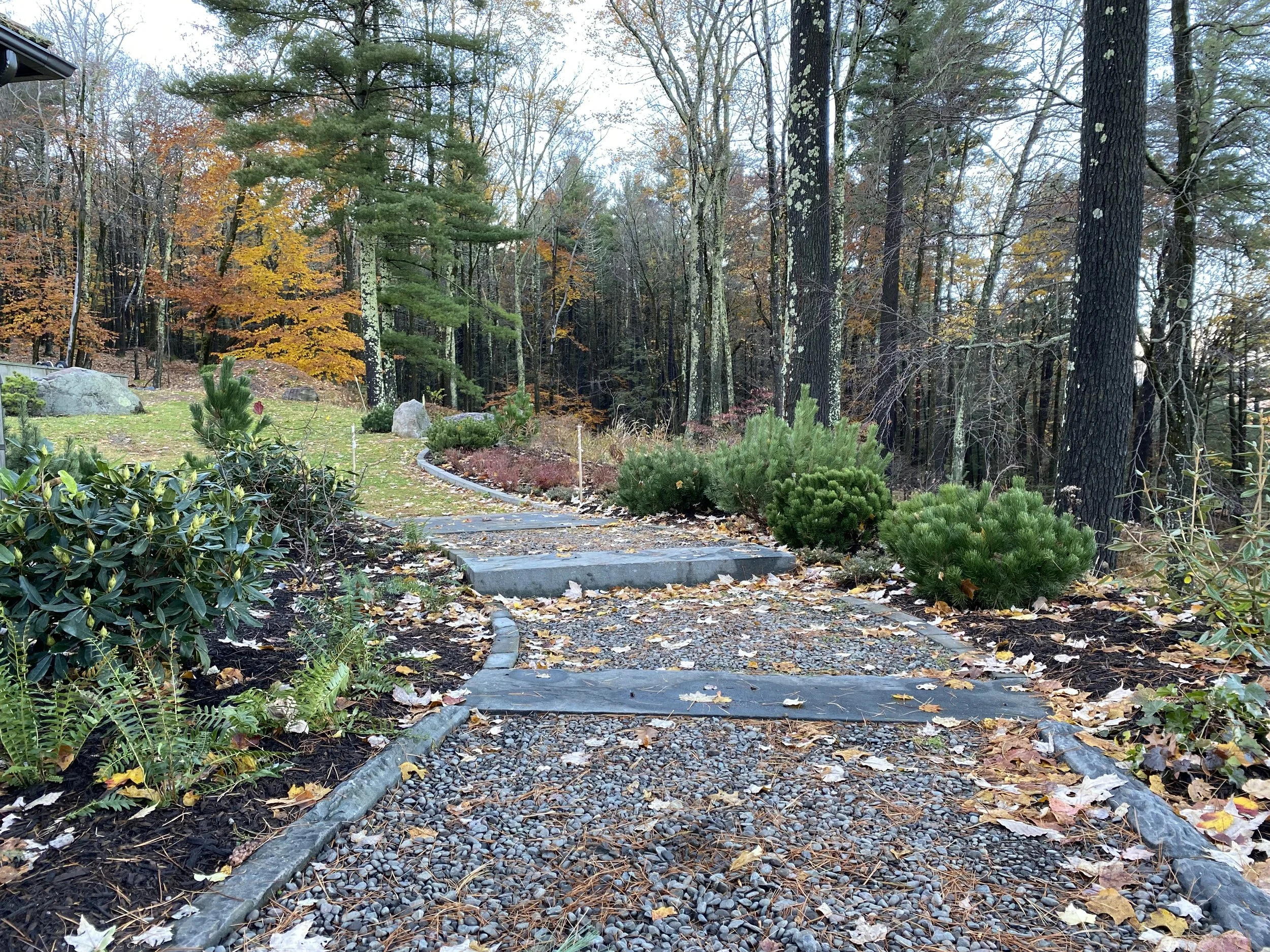 A winding garden pathway made of small gravel and stones, adjacent to green shrubs and autumn leaves, leading into a wooded area with tall trees and fall foliage.