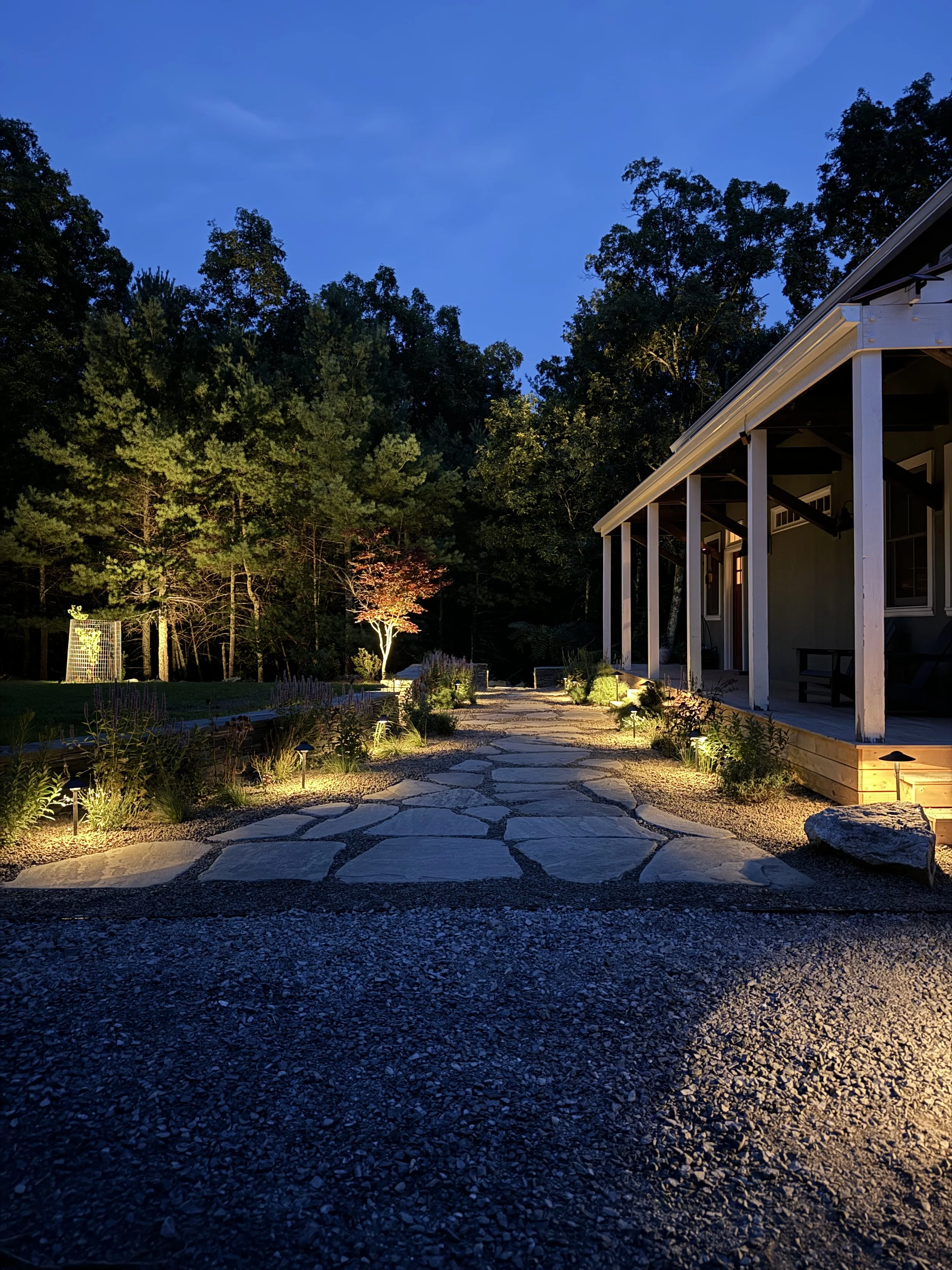 Evening outdoor scene showing a stone pathway leading to a house with porch lighting, surrounded by trees and garden plants under a dark blue sky.