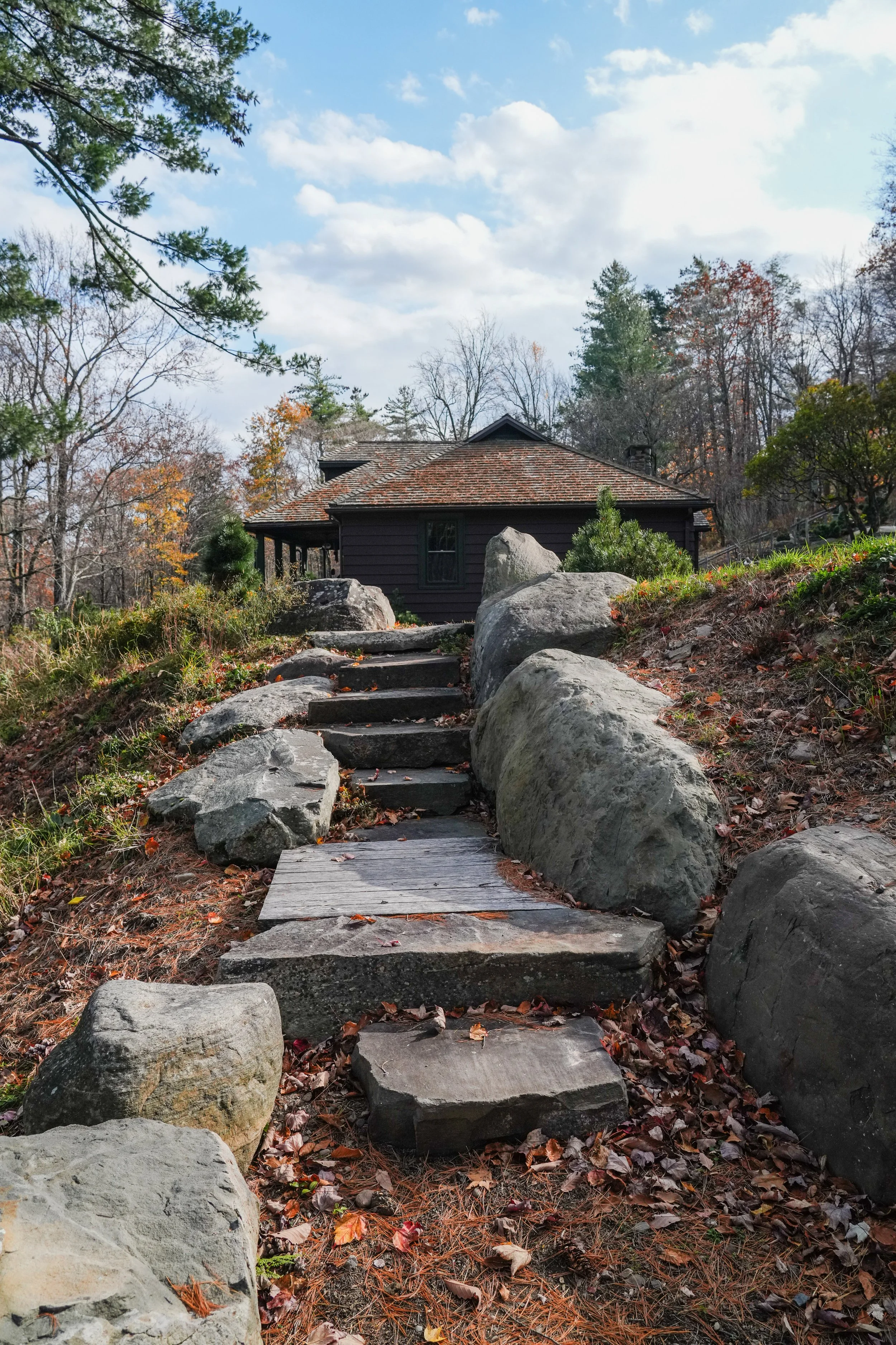 A stone pathway leading up to a dark brown wooden house with a shingled roof, surrounded by trees with autumn foliage under a partly cloudy sky.