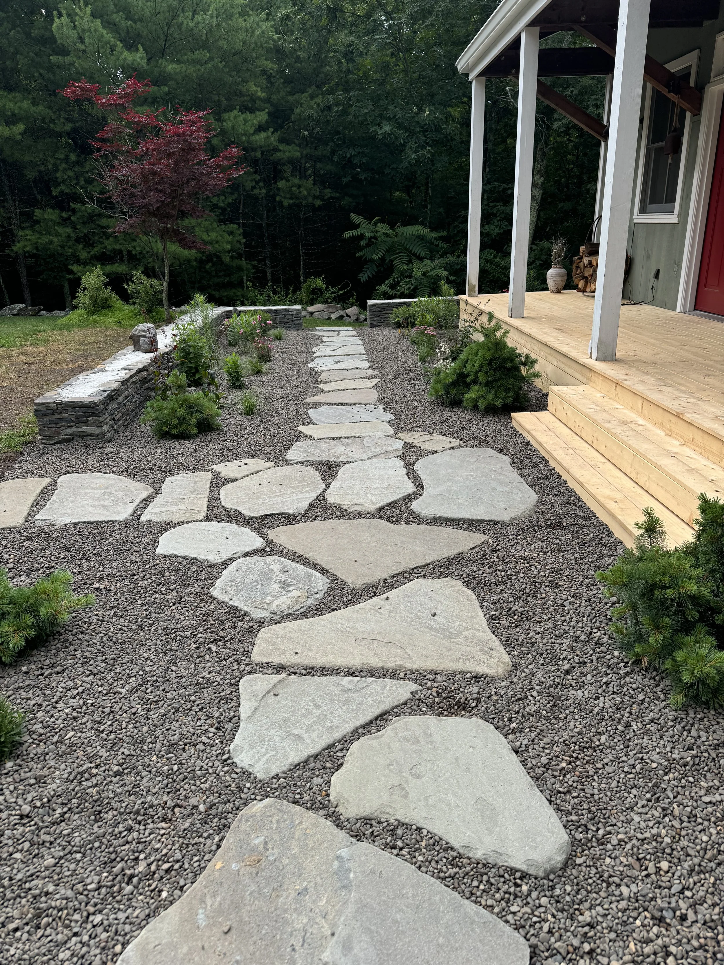 A gravel pathway with large flat stones leading to a wooded area, bordered by small plants and a stone retaining wall on the left, adjacent to a porch with wooden steps and support beams, and lush green trees in the background.