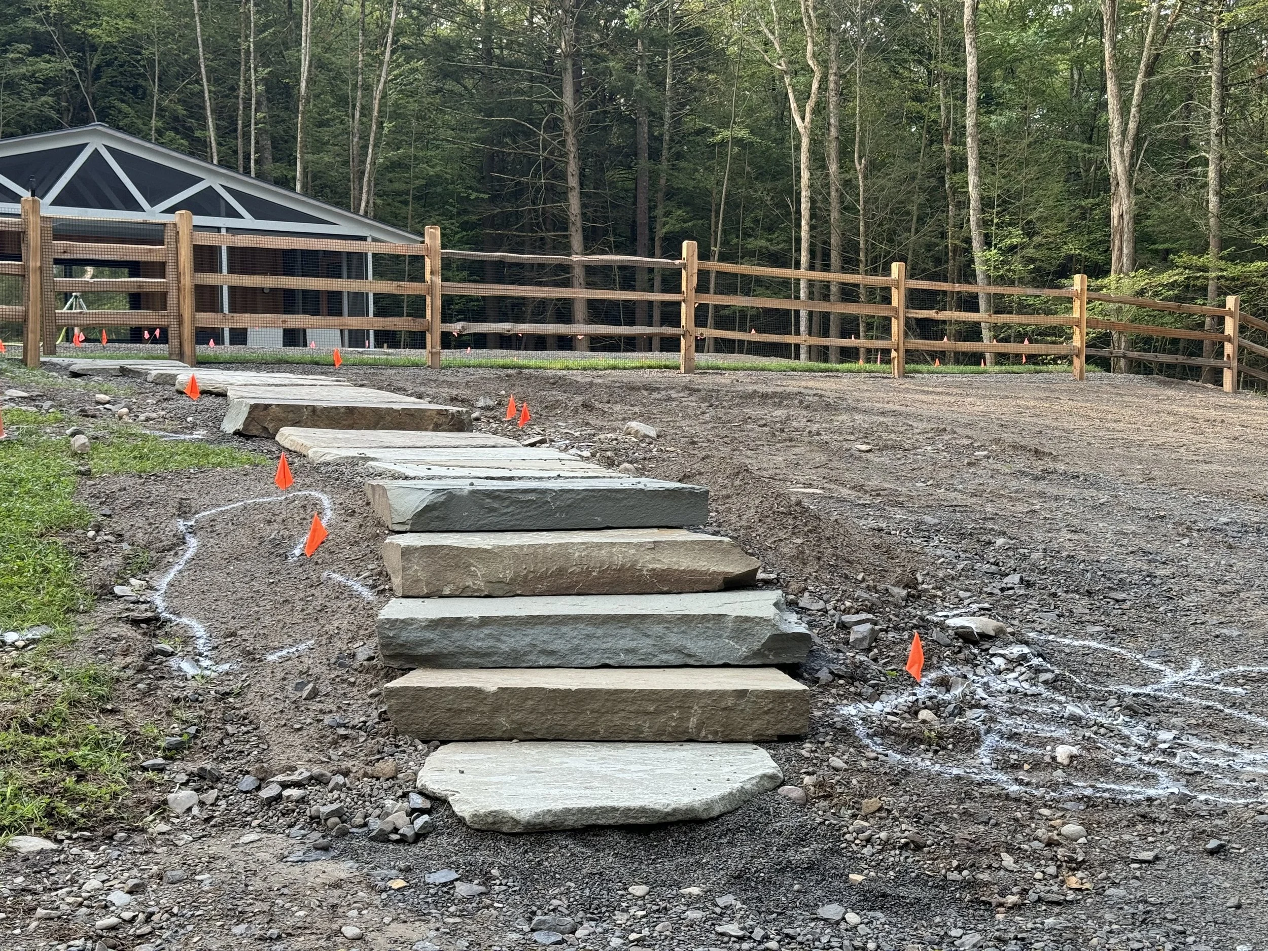 A construction site with a pathway made of concrete steps leading up to a fenced area. The fence is wooden, and there are orange safety flags on the ground indicating the construction zone. A building with a dark roof is visible in the background, su