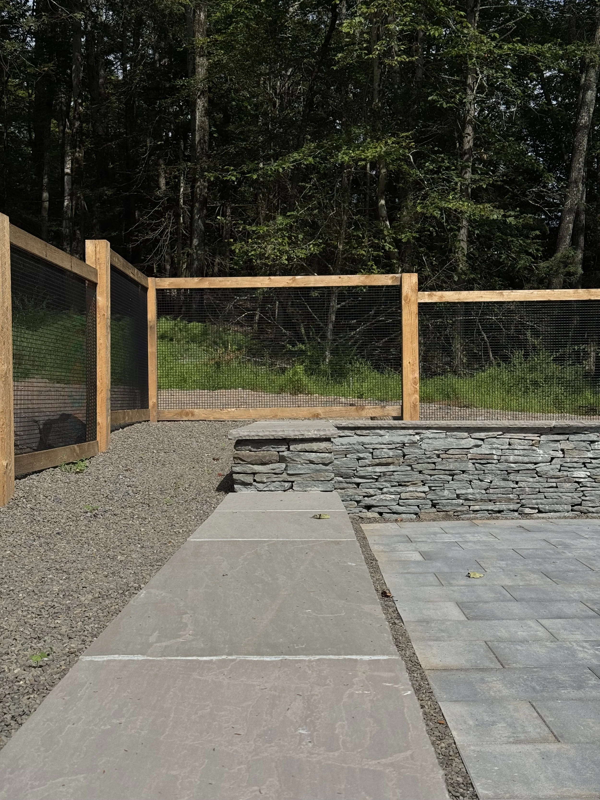Outdoor backyard area with a stone patio, concrete walkway, wooden and wire fence, green grass, and a dense wooded area in the background.