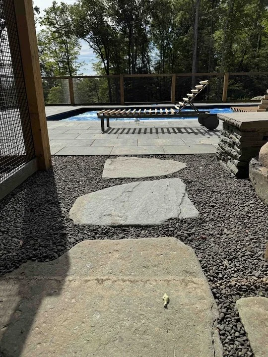 Stone pathway leading to a backyard pool area with lounge chairs and a view of trees and water in the background.