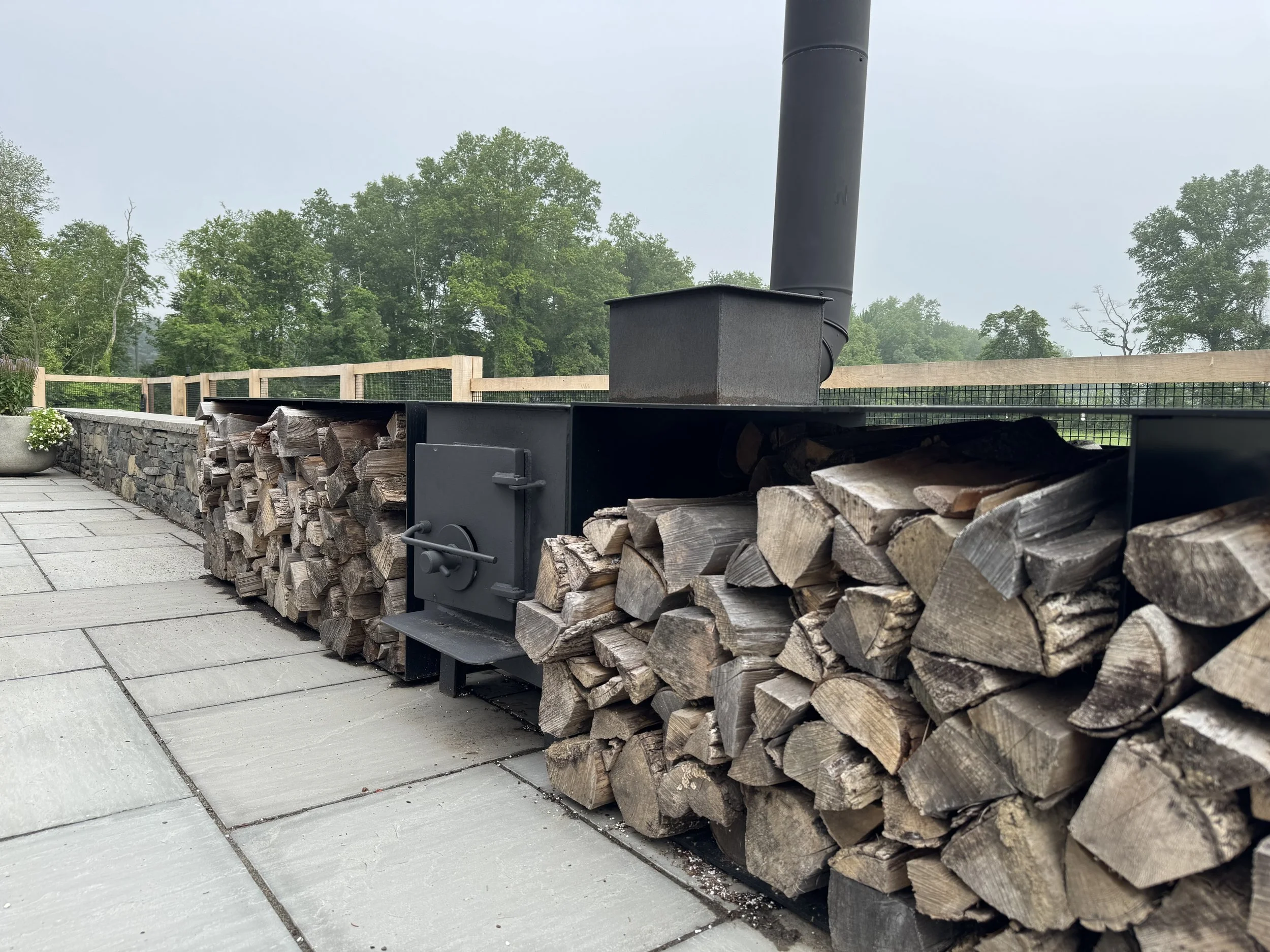 Stacked firewood logs arranged neatly beside a black outdoor stove or fireplace, with a cloudy sky and green trees in the background.