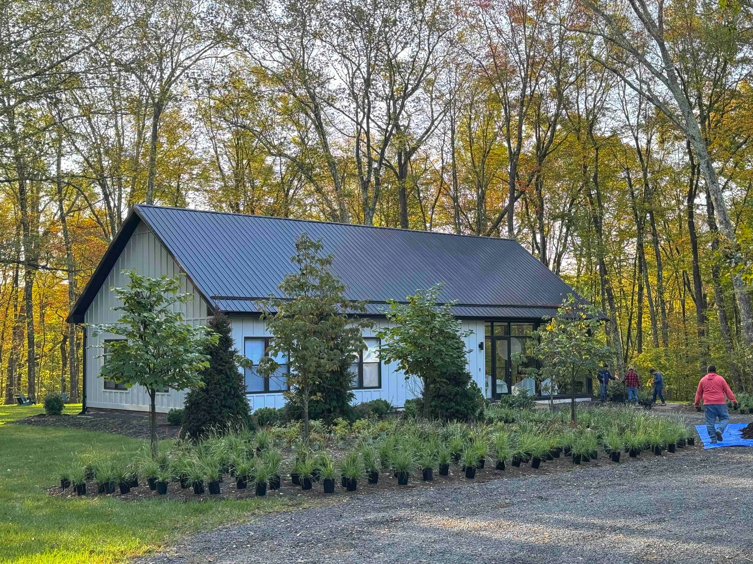 A modern house with a metal roof surrounded by trees and a garden, with people walking nearby.