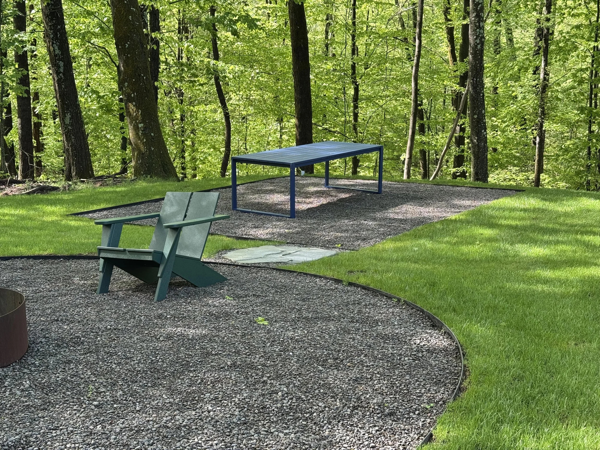 A backyard with green grass, a gravel patio area with a rectangular blue table, two green chairs, and a wooded area with trees in the background.