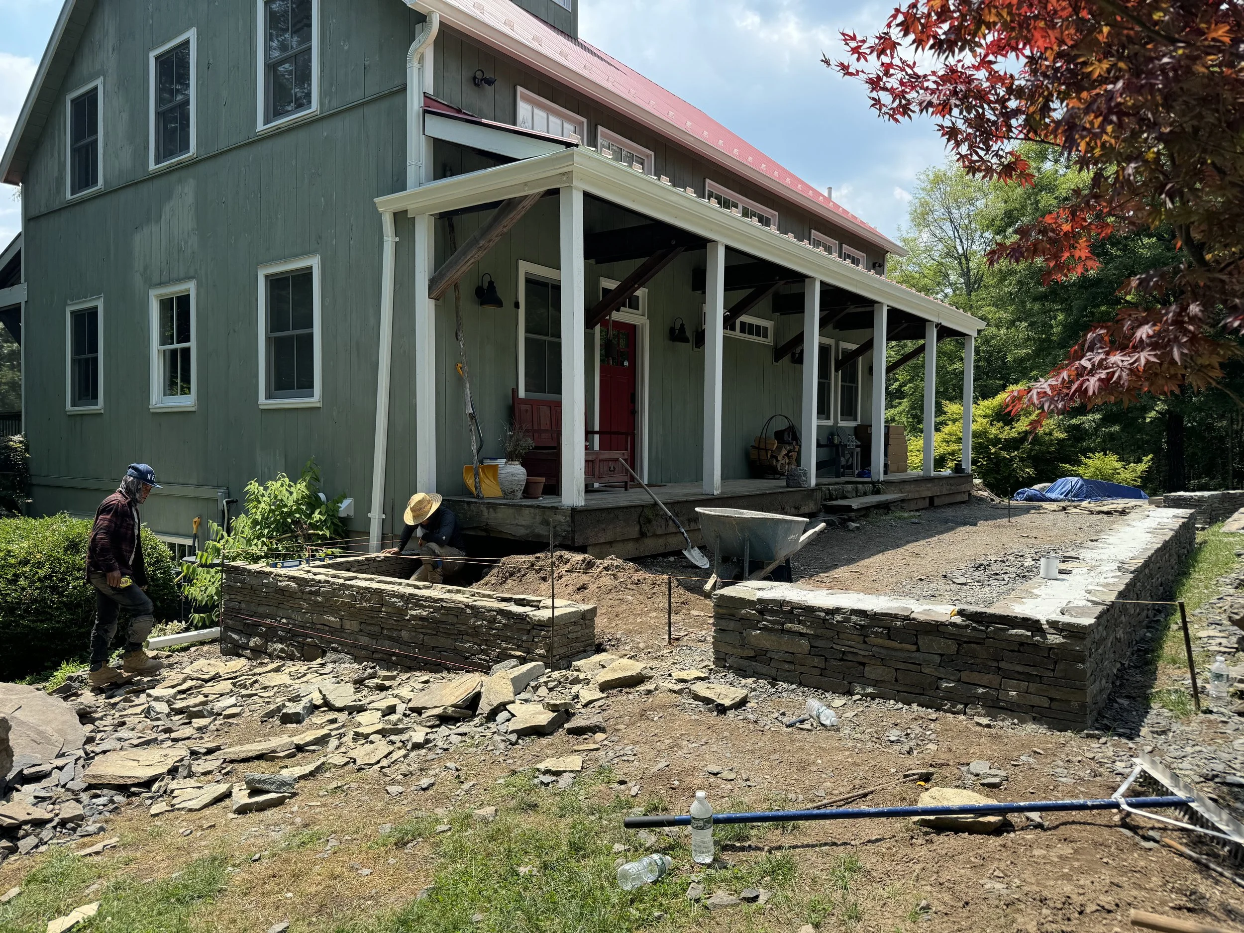 Construction workers building a stone wall in the yard of a green house with a porch and red roof, surrounded by trees and construction tools.