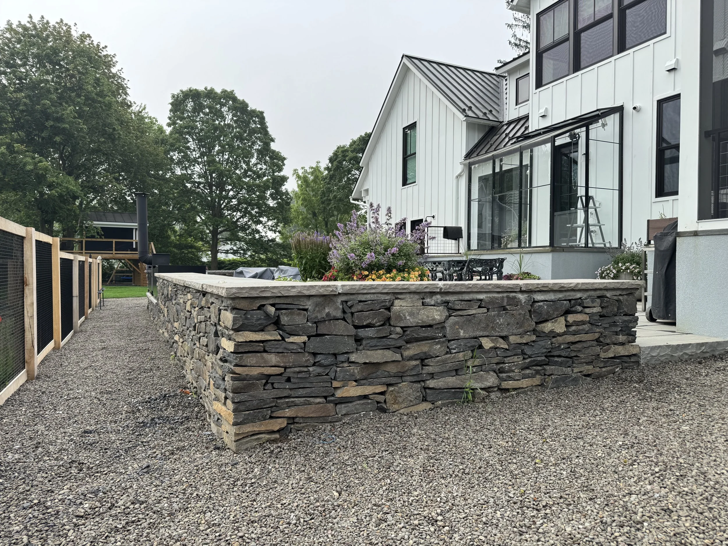 A modern two-story house with white vertical siding and large windows, surrounded by a landscaped yard with a stone wall, purple flowers, and outdoor seating, with trees in the background.