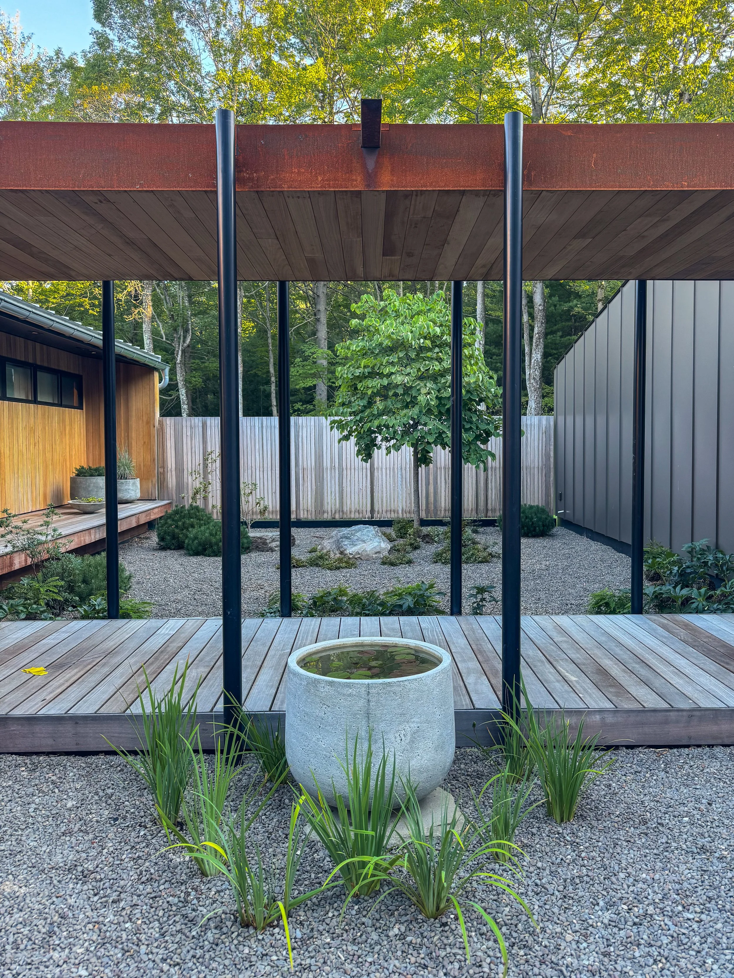 A modern outdoor garden with a pergola, a small pond in a stone planter, and various green plants and shrubs.