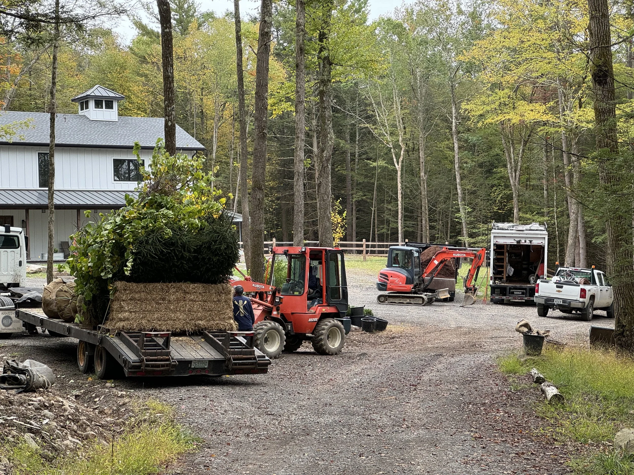 A construction site in a wooded area with a white house, construction vehicles including a red skid steer loader and a truck, and construction materials and equipment scattered around.