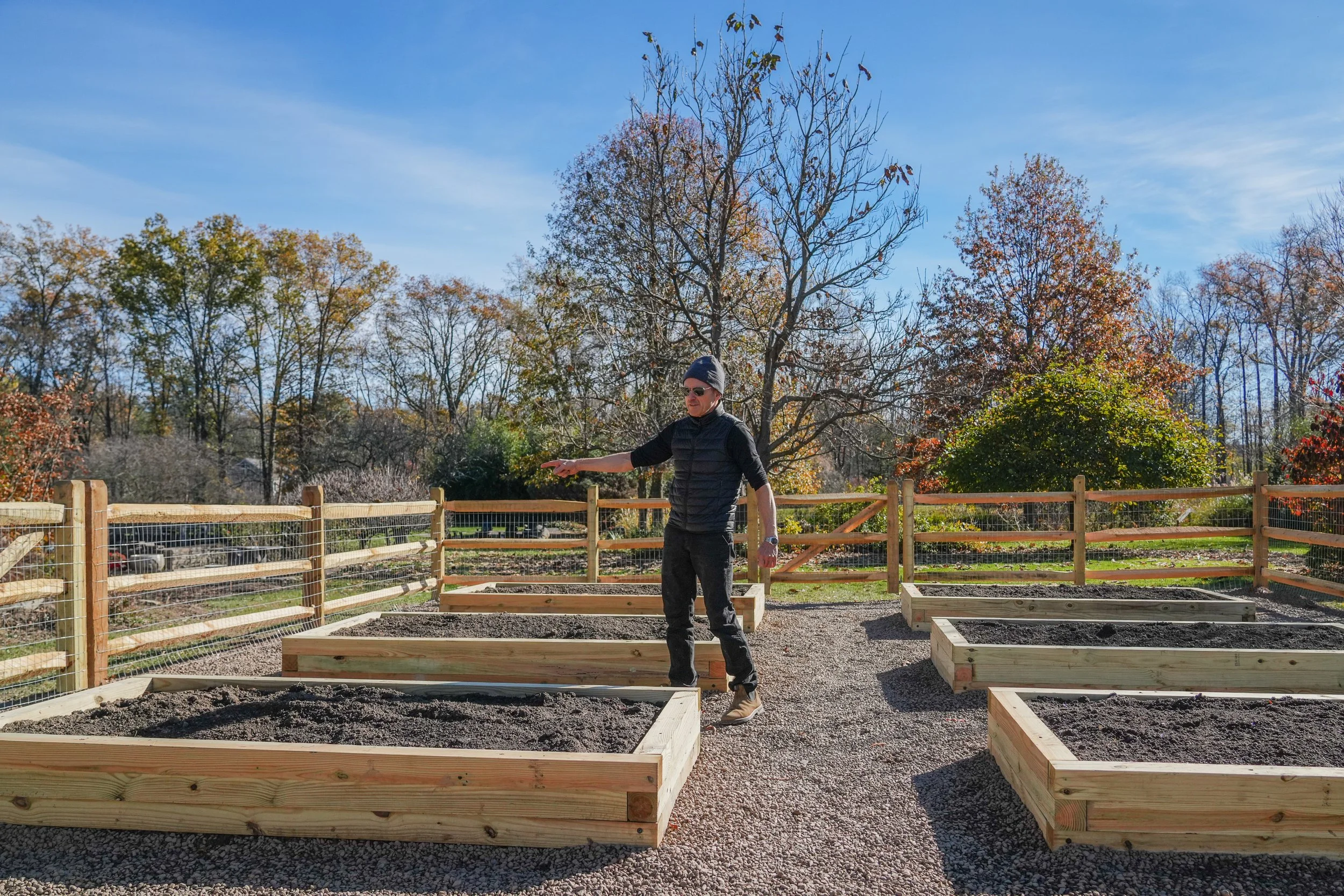 A man wearing sunglasses, a beanie, and a black vest is standing in a garden with square wooden planting beds filled with soil. He is pointing to the right. The garden is surrounded by a wooden fence, with trees displaying autumn foliage and a clear 