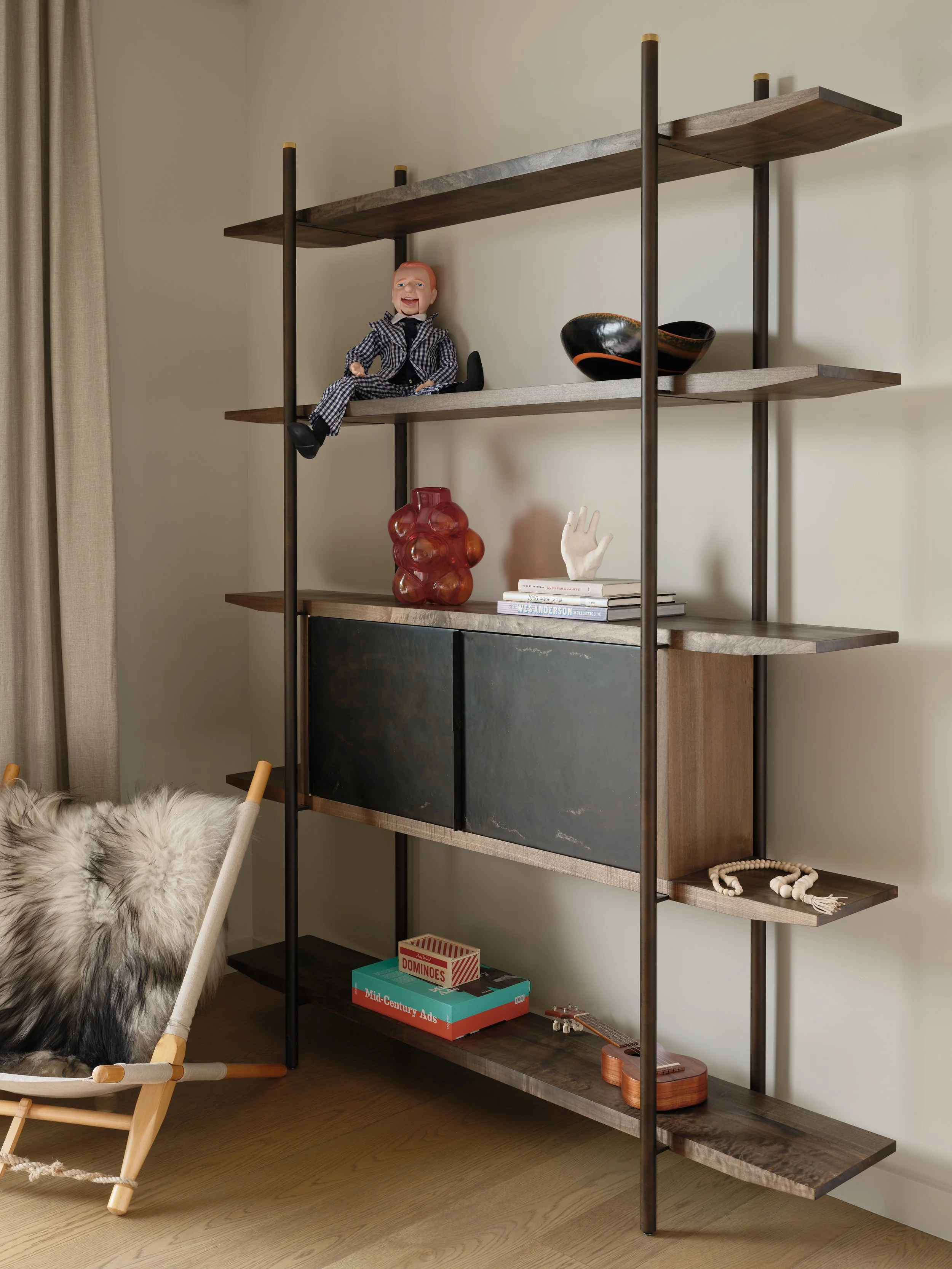 A modern wooden and metal bookshelf with decorative items, a doll, vases, books, a string of beads, a guitar, and a box of dominoes, positioned next to a cozy chair with a fur throw.